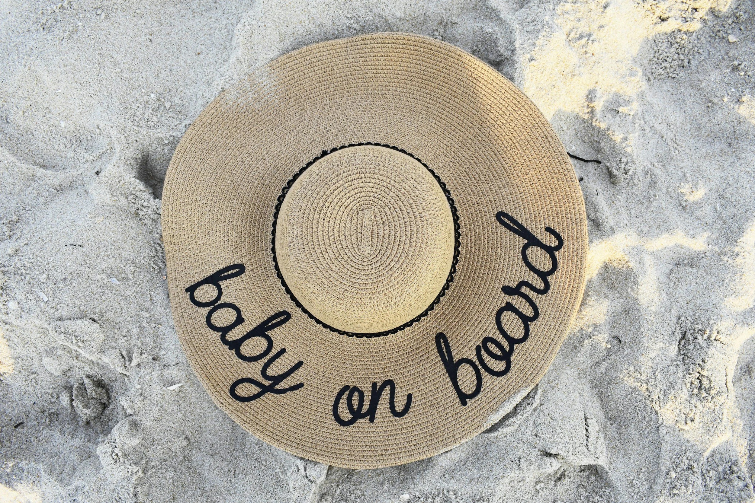 A tan woven sunhat with black cursive text that reads "baby on board" lying on sandy beach ground.