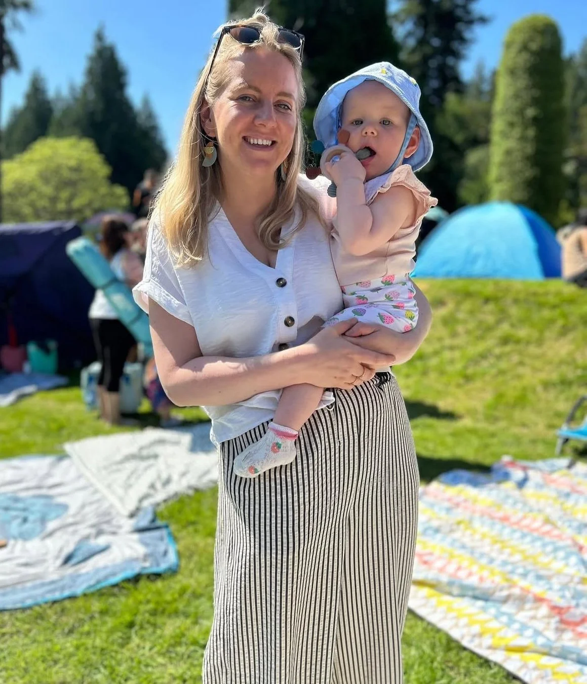 A woman holding a young girl outdoors on a sunny day with tents and people in the background.