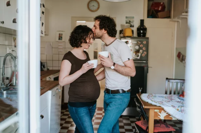 A couple sharing a kiss in the kitchen, holding cups, with a woman wearing a brown shirt and the man in a white t-shirt and jeans.