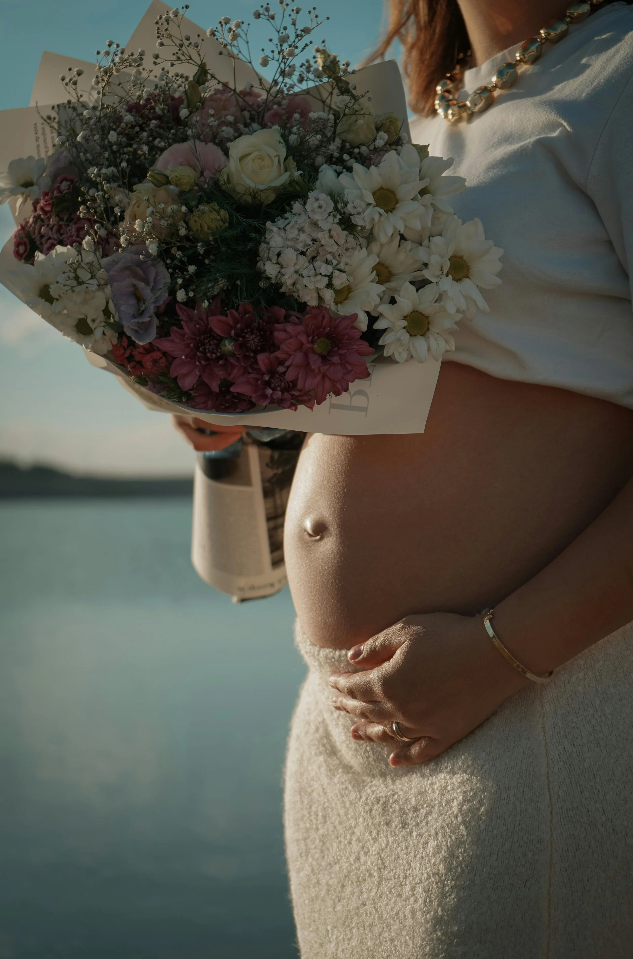 pregnant tummy exposed in the evening sunlight. woman is holding some beautiful flowers with her hand on her stomach