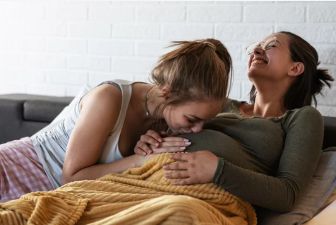 Two women smiling, one woman is pregnant, one woman is kissing her belly while lying on a bed.