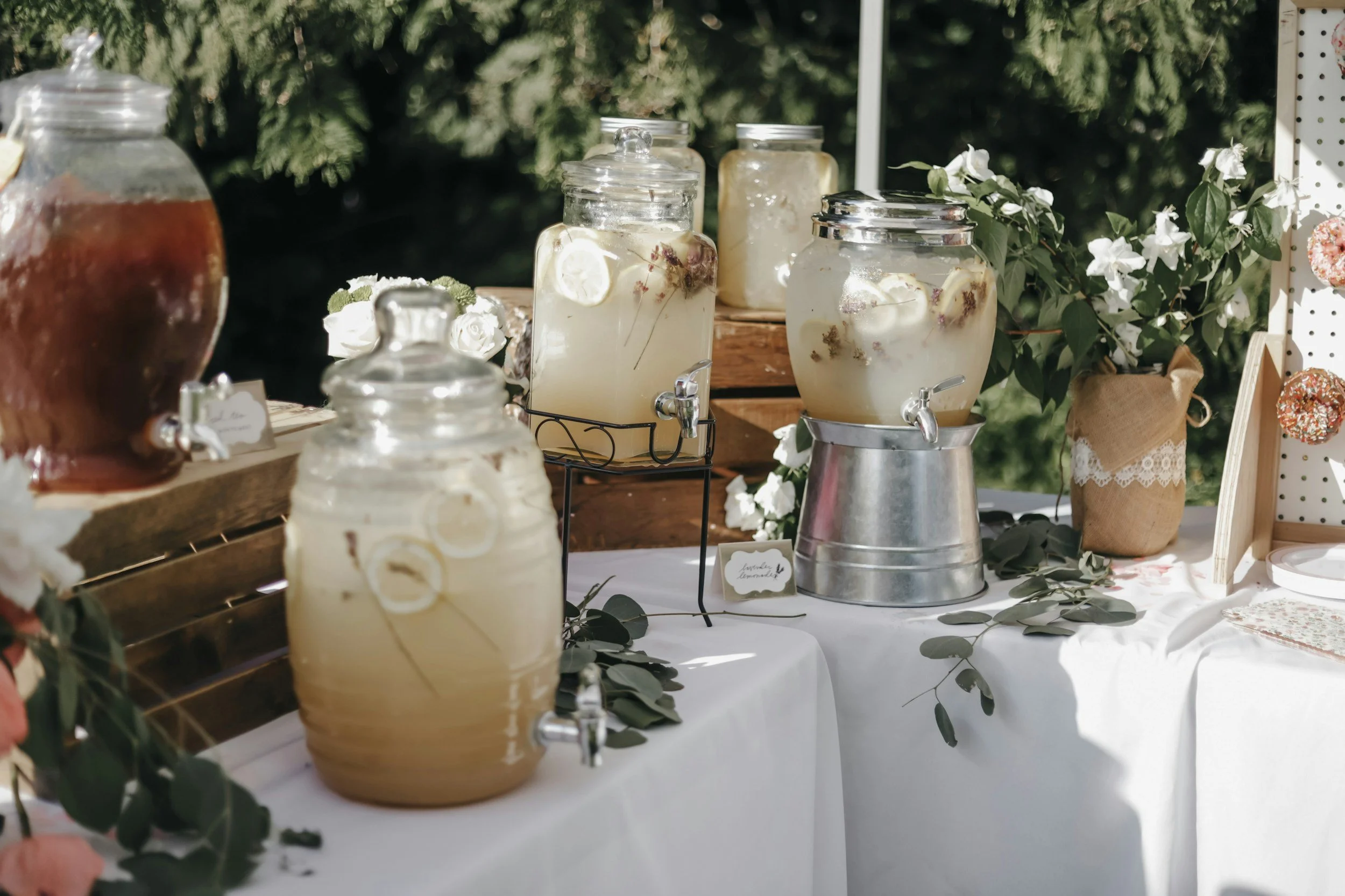Jars of flavored lemonade and iced tea on a white tablecloth at an outdoor event, decorated with green foliage and flowers.