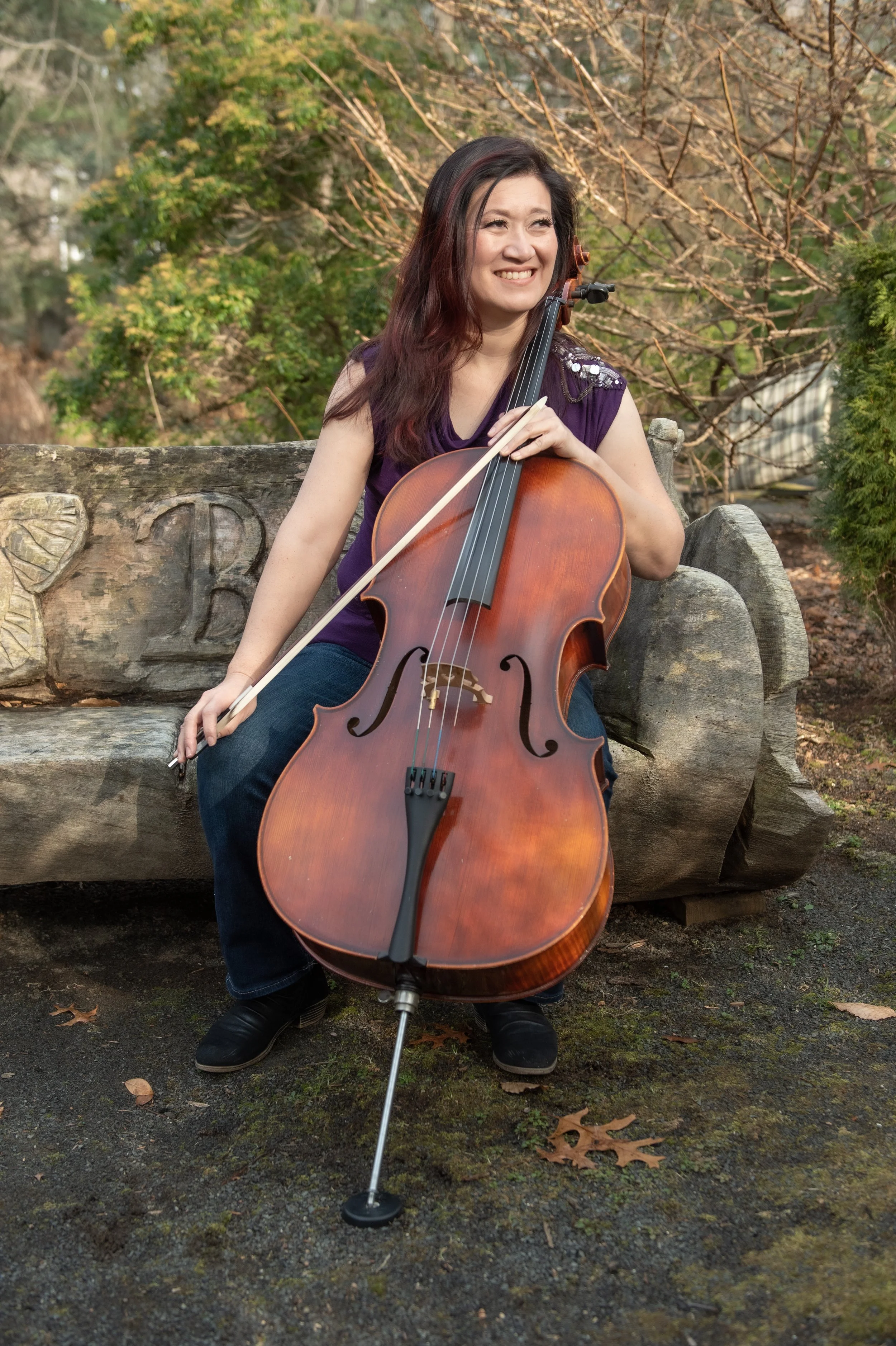 A woman sitting on a wooden bench outdoors, smiling, and holding a cello with her left hand while her right hand holds a bow.