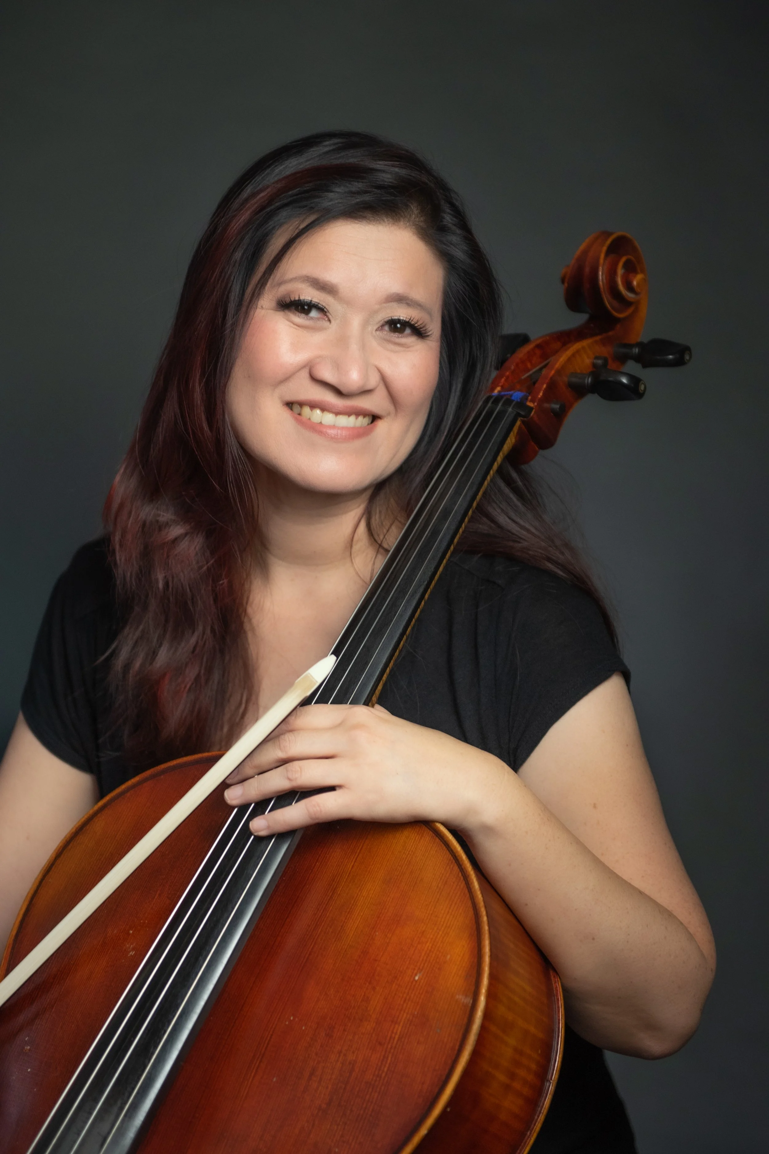 A woman with long dark hair holding a cello and smiling at the camera.