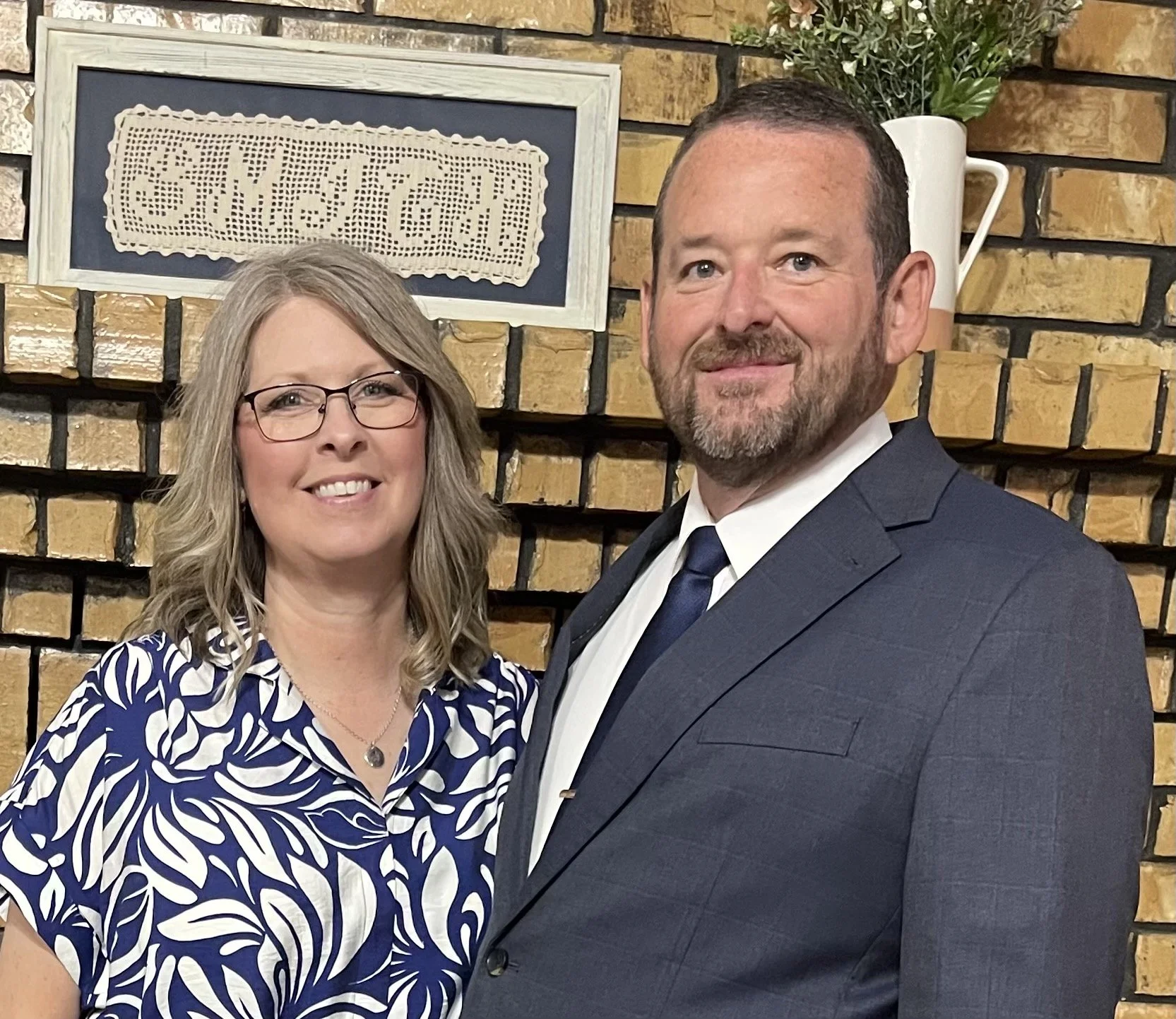 A smiling couple posed together indoors in front of a brick wall. The woman has shoulder-length blonde hair, glasses, and is wearing a blue and white patterned blouse. The man has short dark hair, a beard, and is dressed in a dark suit with a white shirt and a blue tie.