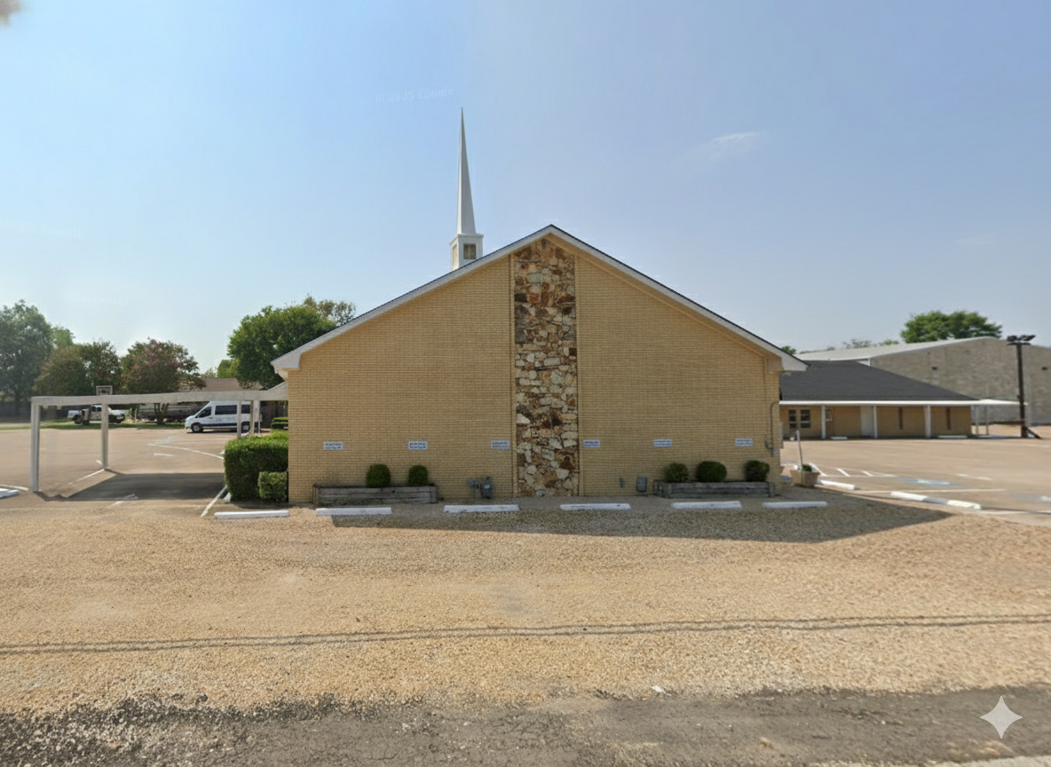 a church building with a brick exterior and a stone vertical strip in the middle, with a tall white steeple, a parking lot surrounding it, and some bushes in front.