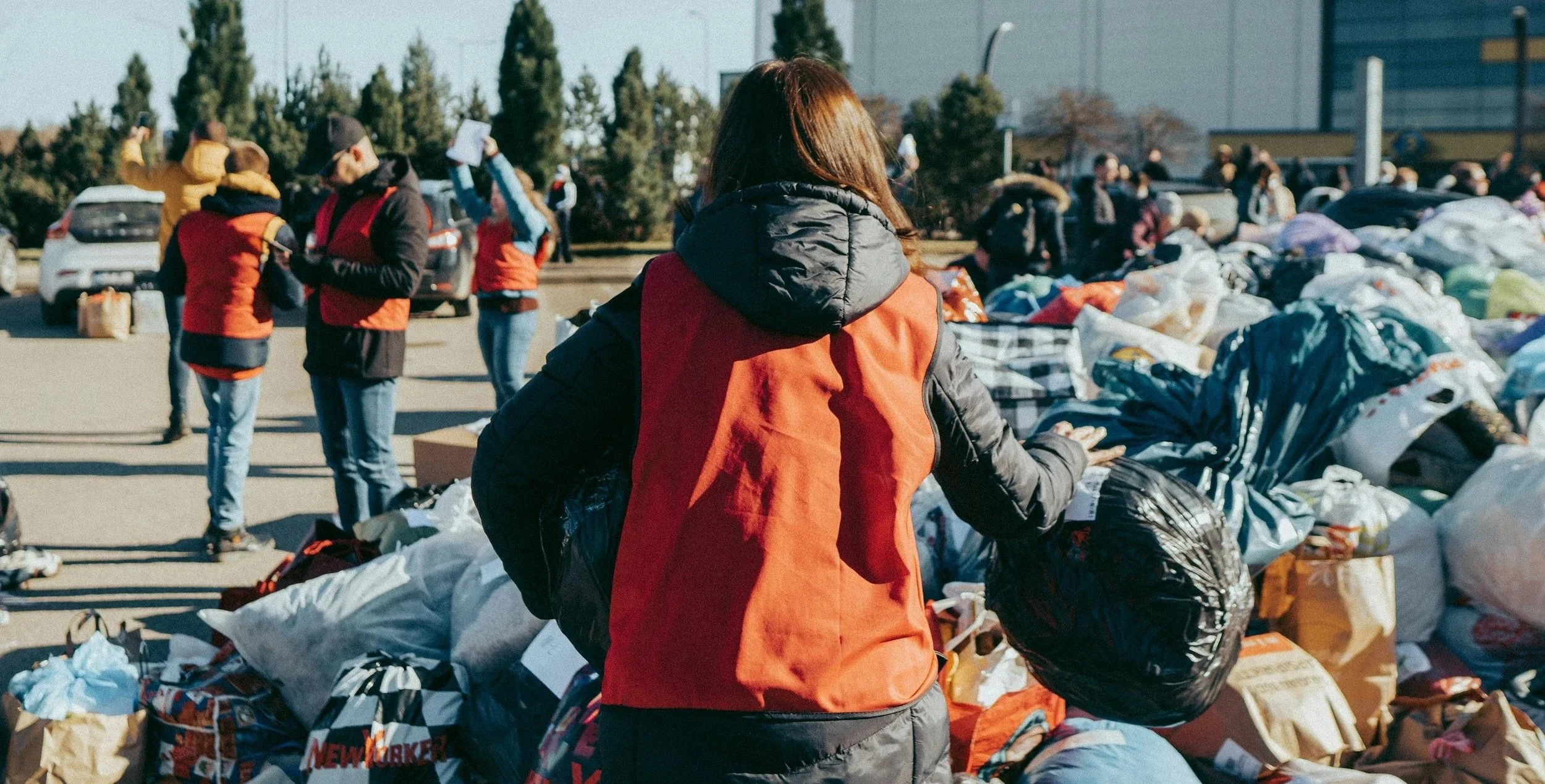 Person in orange jacket and black hoodie organizing large trash bags among piles of donated clothing and supplies outdoors, with other people in the background.