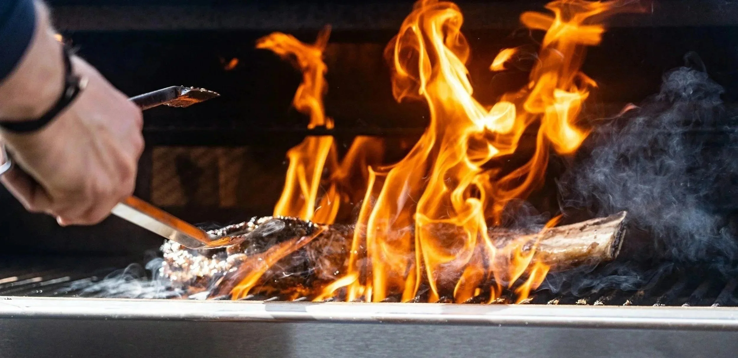 Person tending to a BBQ grill with flames and smoke rising from burning logs.