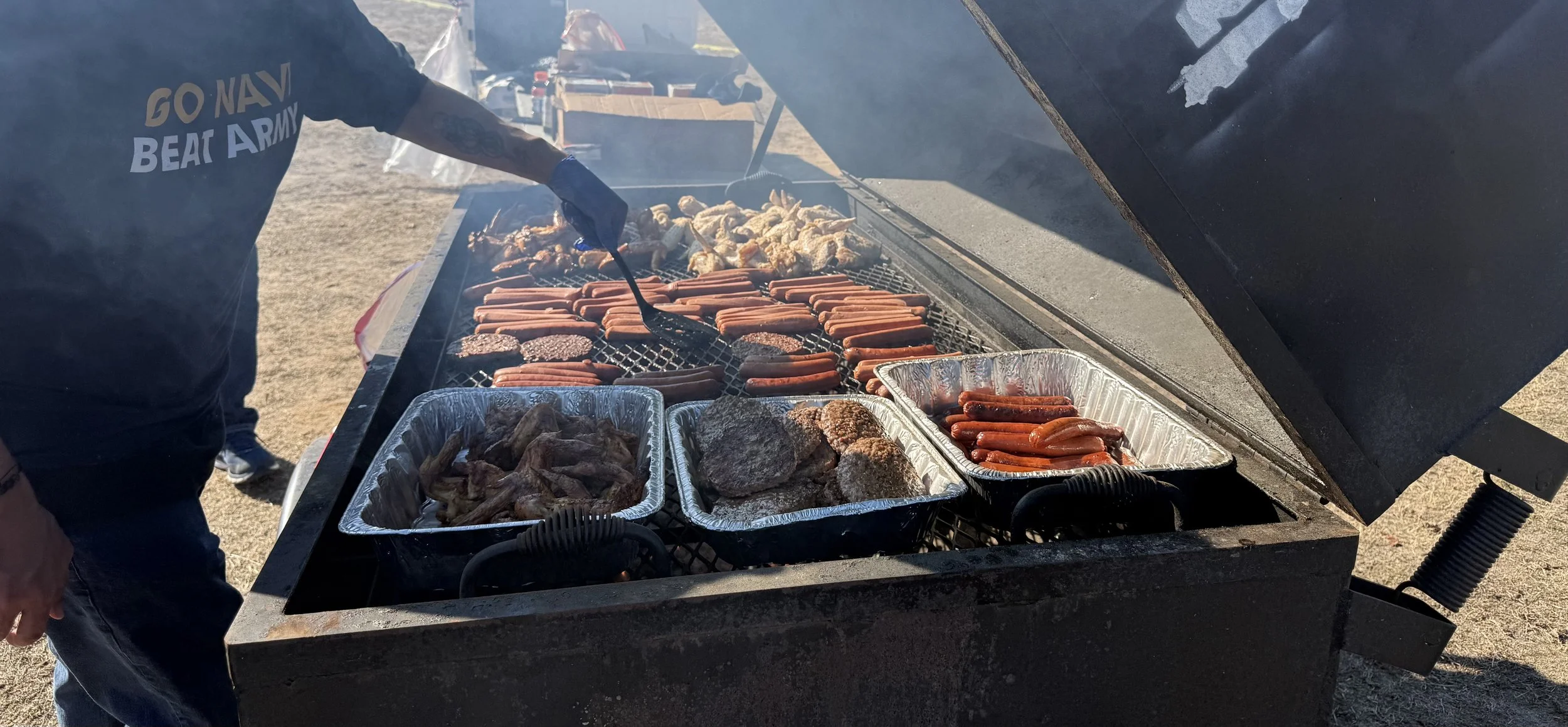 Grill with hot dogs, chicken wings, and burgers cooking, with aluminum trays of cooked meat on the side, outdoors.