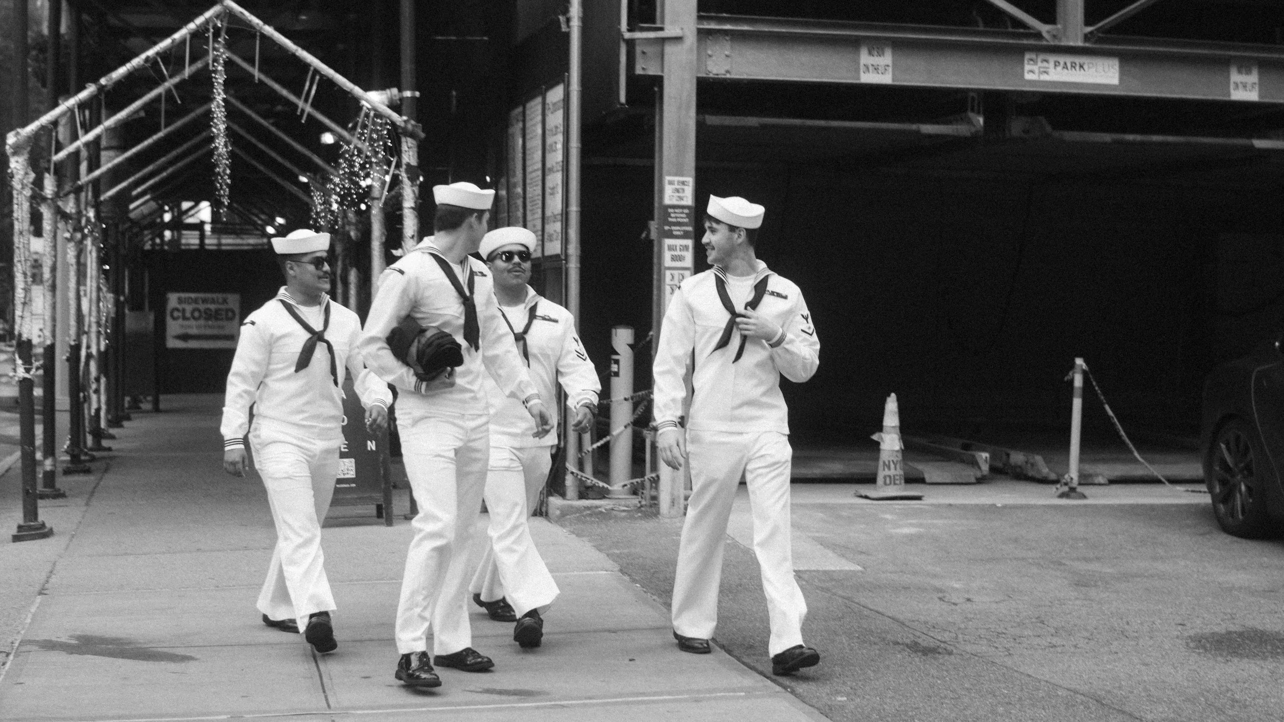 Four men dressed in naval uniforms walking together on city sidewalk near scaffolding and a parking garage, with holiday decorations hanging overhead.