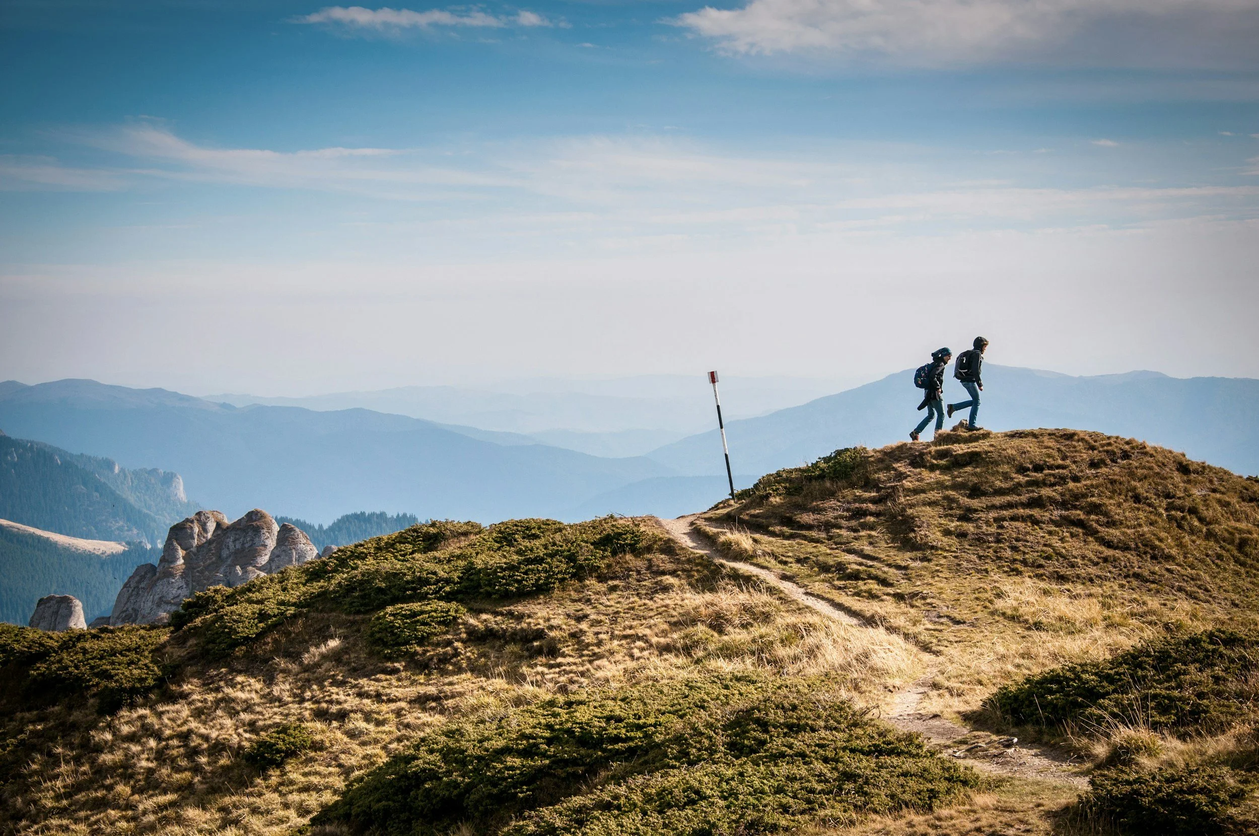 Two hikers walking along a mountain trail with scenic mountain landscape in the background.