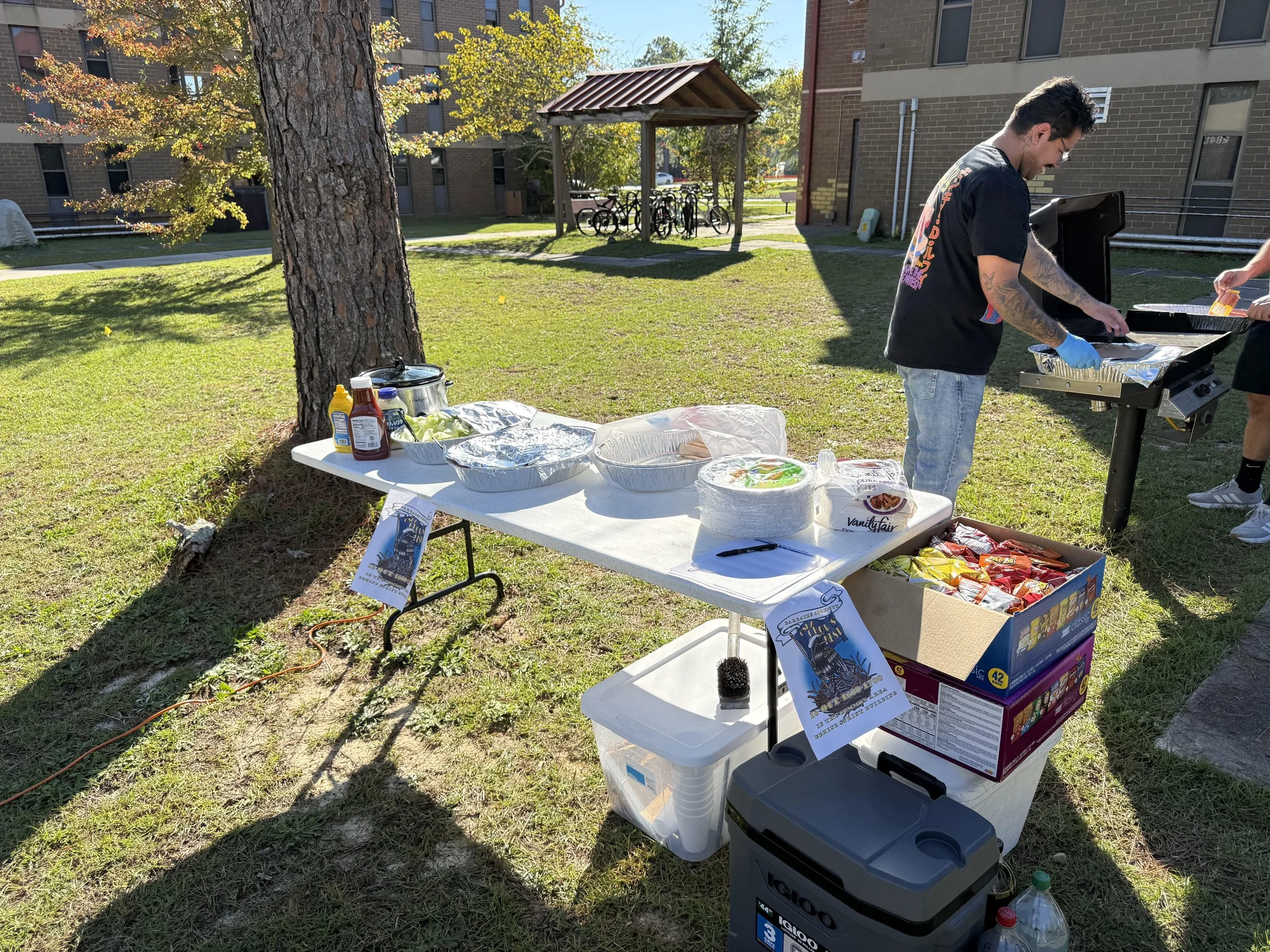 Outdoor barbecue and food setup with condiments, chips, and foil-wrapped food on a table, a man grilling, in a grassy area near apartment buildings on a sunny day.