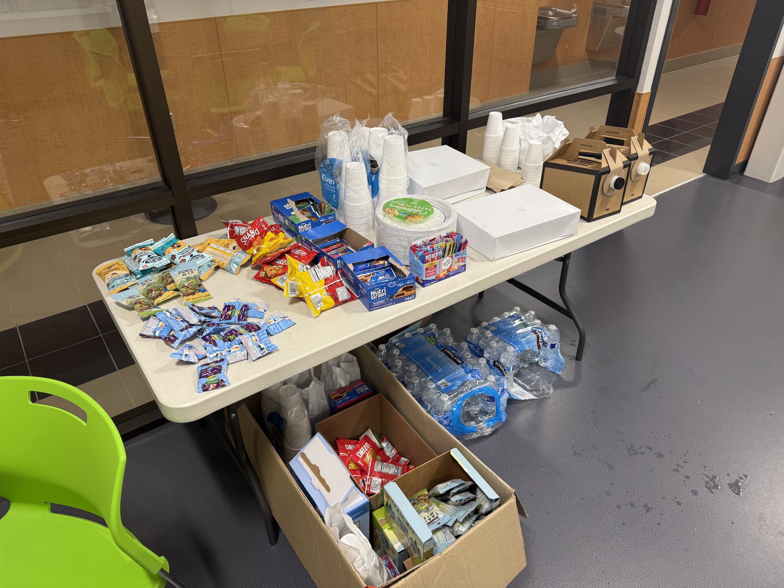 Table with assorted snacks, bottled water, disposable cups, and cardboard boxes in a room with a glass wall.