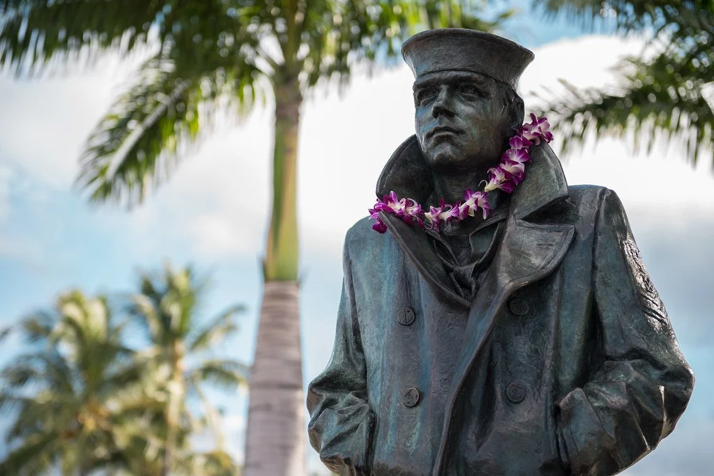Bronze statue of a man wearing a military cap and coat, with a purple flower lei around his neck, set against a background of palm trees and partly cloudy sky.
