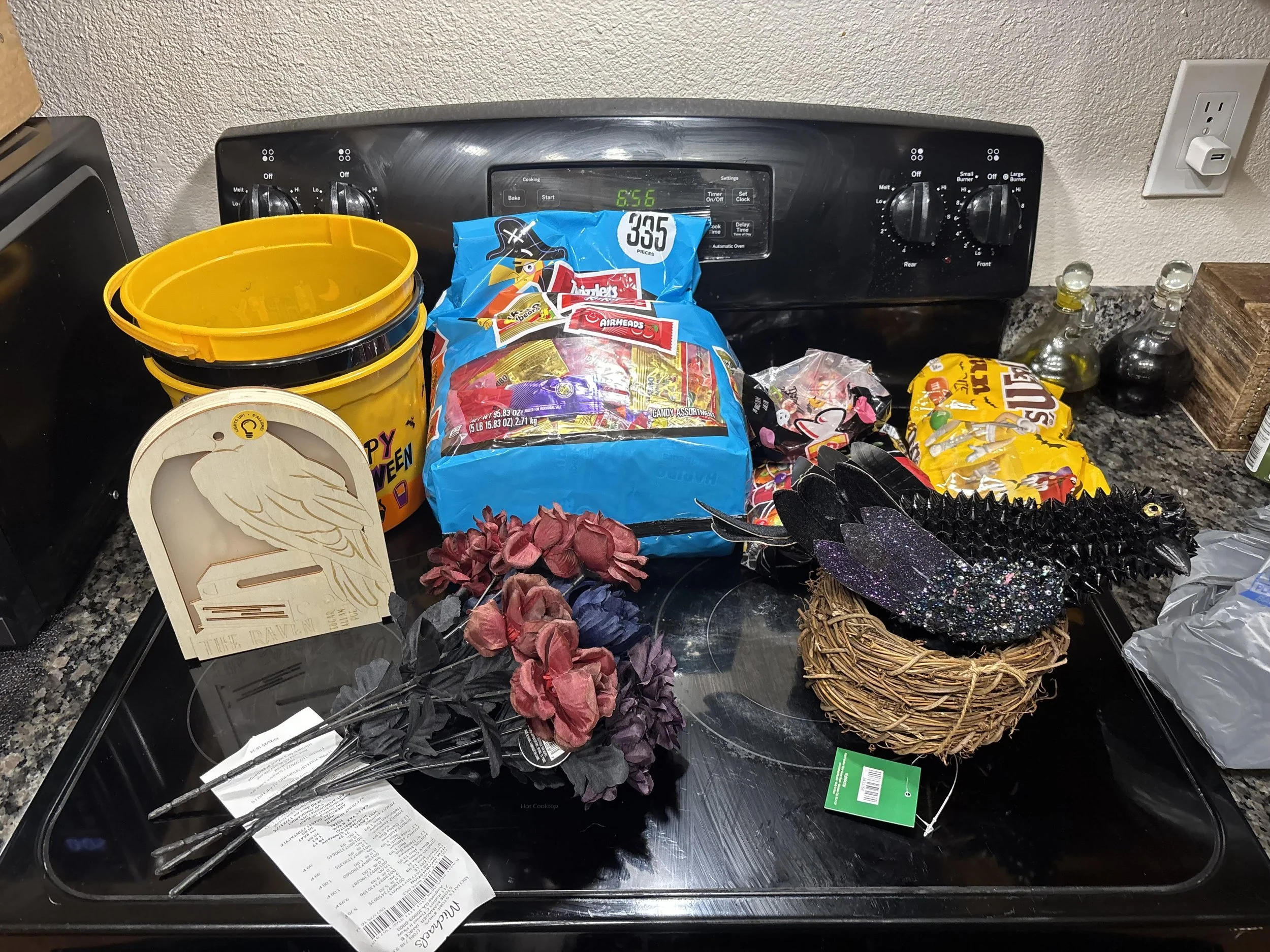 Cluttered kitchen stovetop with Halloween decorations, snack bags, yellow buckets, a swan-themed sign, and condiments.