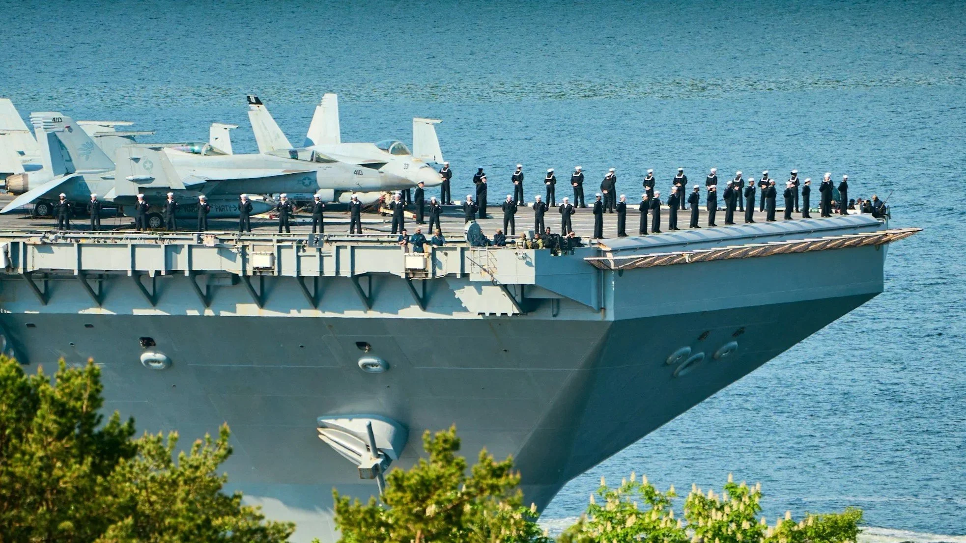 U.S. Navy aircraft carrier deck with Jet fighter jets and sailors standing at attention, with green trees in the foreground and water in the background.
