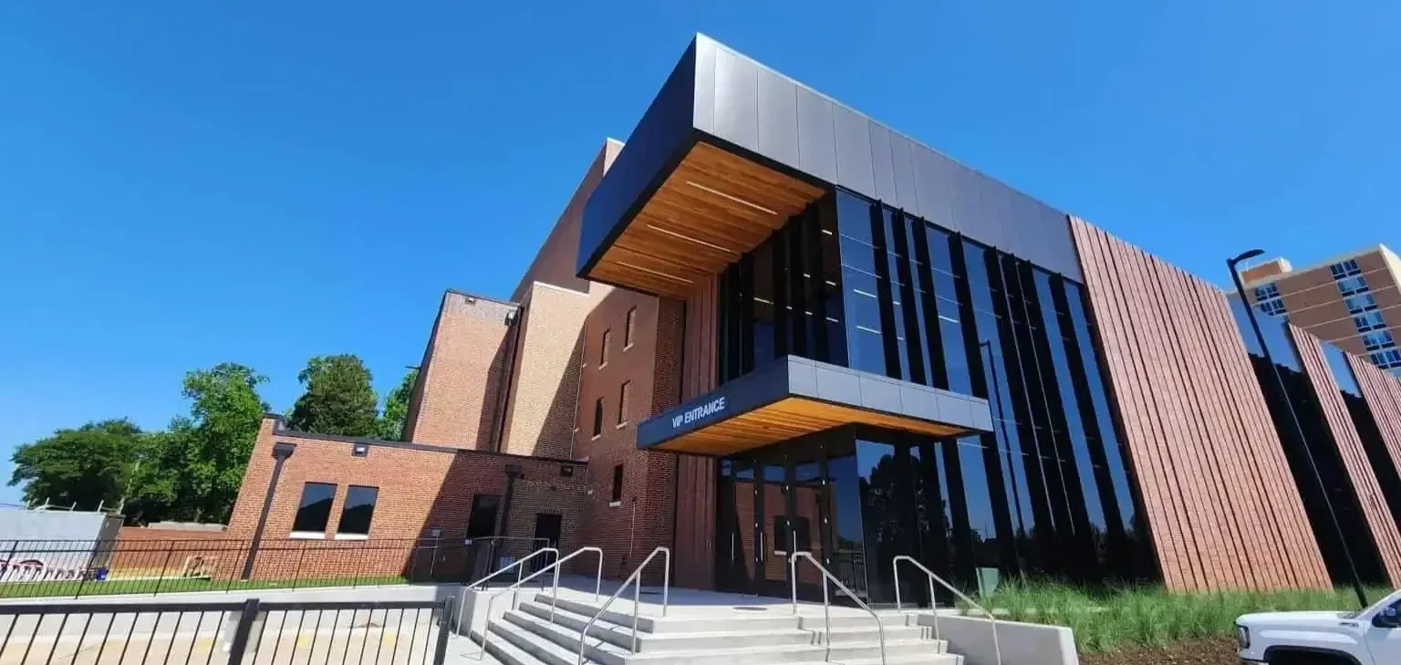 Modern multi-story building with a mix of brick and black glass exterior, featuring a VIP entrance with stairs and railing, under a clear blue sky.