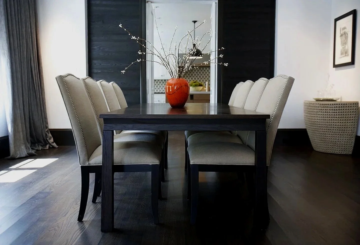 A modern dining room with a dark wooden table, beige upholstered chairs, an orange vase with white flowering branches, dark hardwood floors, a white wall with black trim, a large window with dark curtains, and framed artwork.