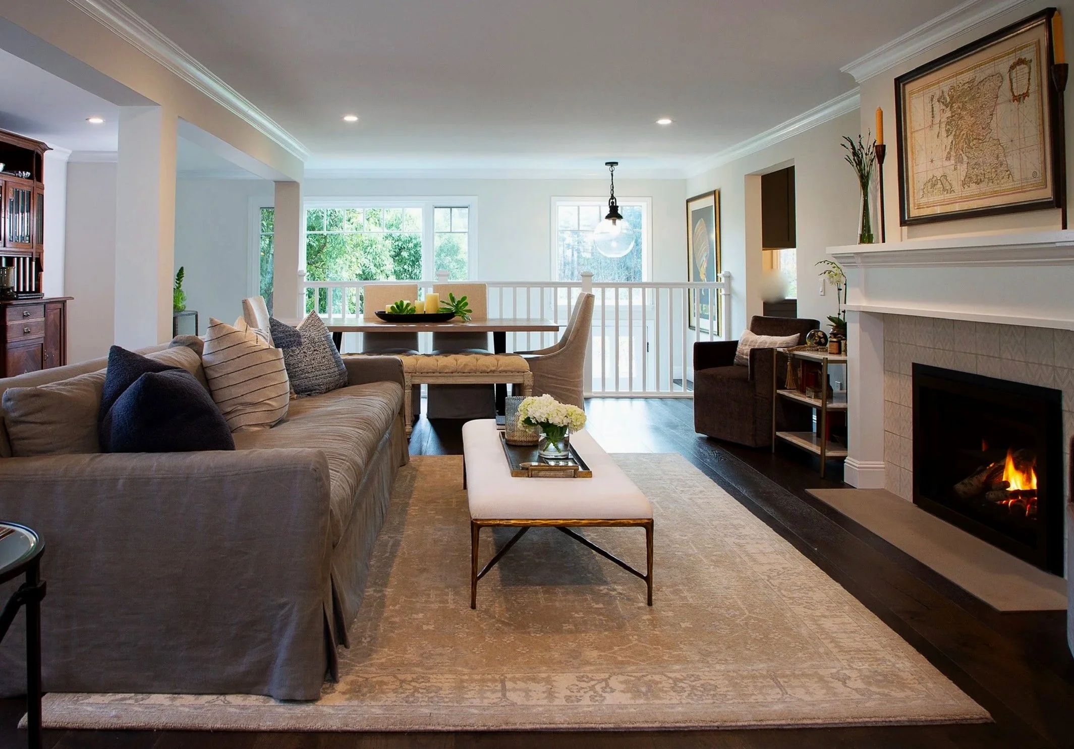 Living room with beige sofa, fireplace, glass-top coffee table with flowers, and dining area with table and chairs, bright windows with greenery outside.
