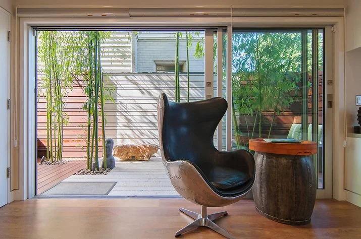 Modern living room with a black, high-back armchair facing an outdoor deck with plants and bamboo, through sliding glass doors.