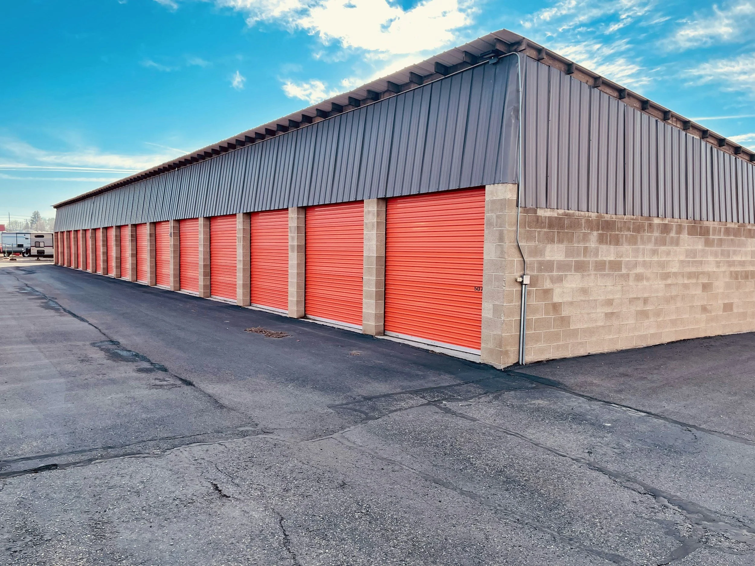A storage unit facility with orange roll-up doors, a gray metal roof, and beige brick walls, under a partly cloudy blue sky.