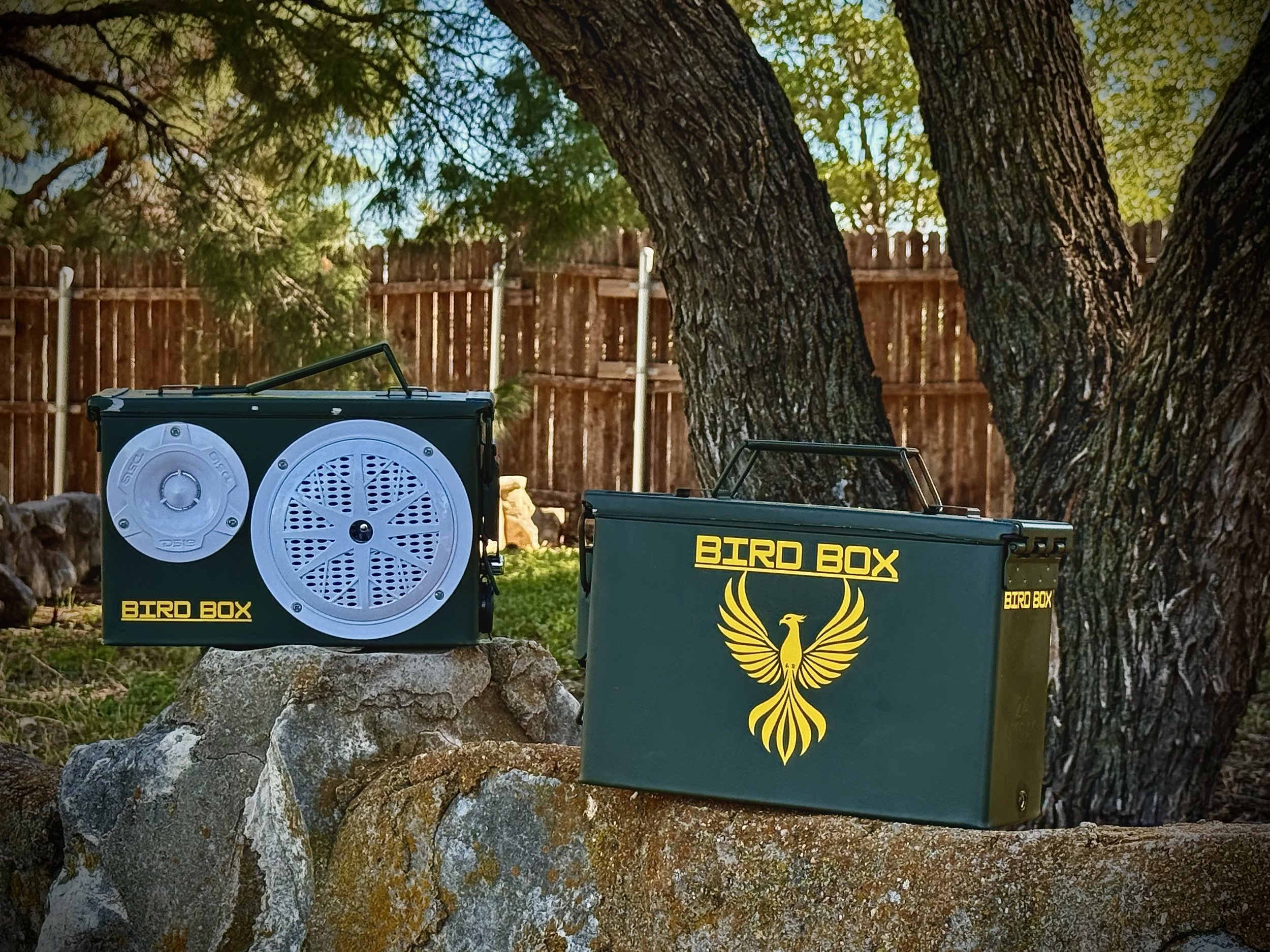 Two green bird boxes with yellow logos and text, one on a rock and one on the ground, in a backyard with a wooden fence and large trees in the background.