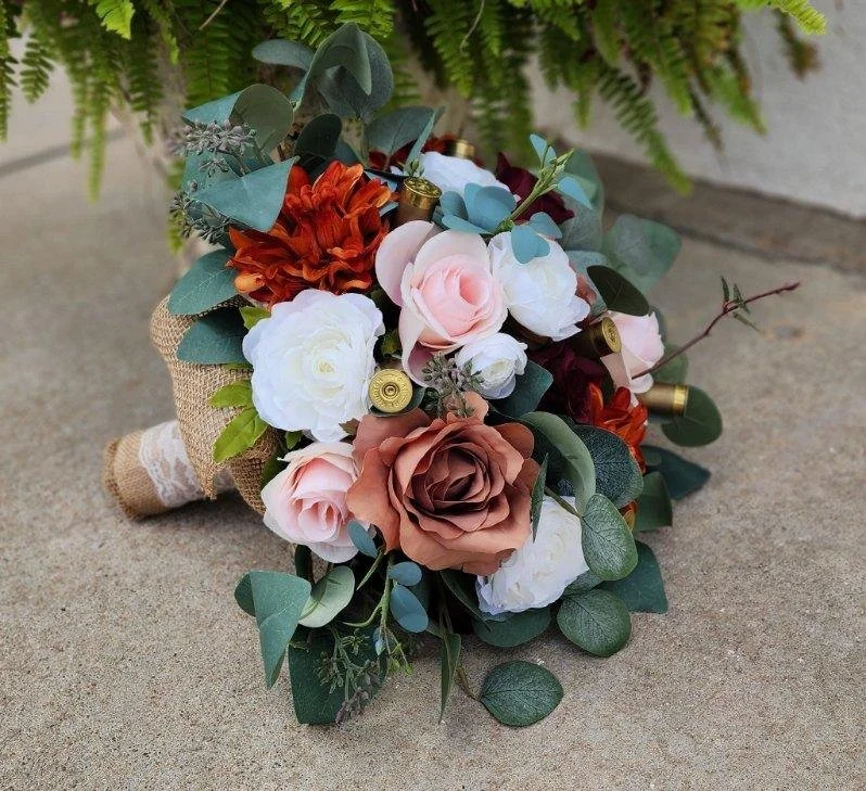 A bouquet of mixed flowers including roses and greenery, with shotgun shells incorporated, resting on a concrete surface with a fern plant in the background.