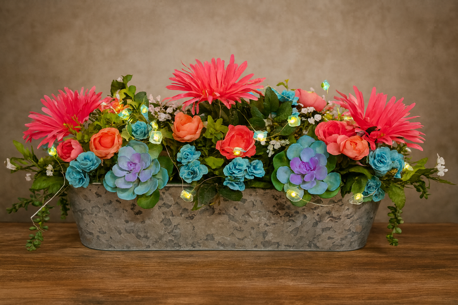A floral arrangement with pink, orange, and blue flowers in a gray metal container, decorated with small string lights, on a wooden surface against a neutral background.