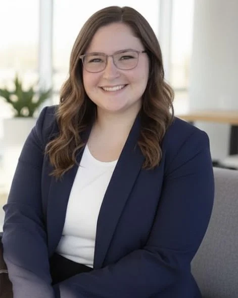 A woman with long wavy brown hair, glasses, wearing a navy blazer over a white top, smiling in a bright office setting.