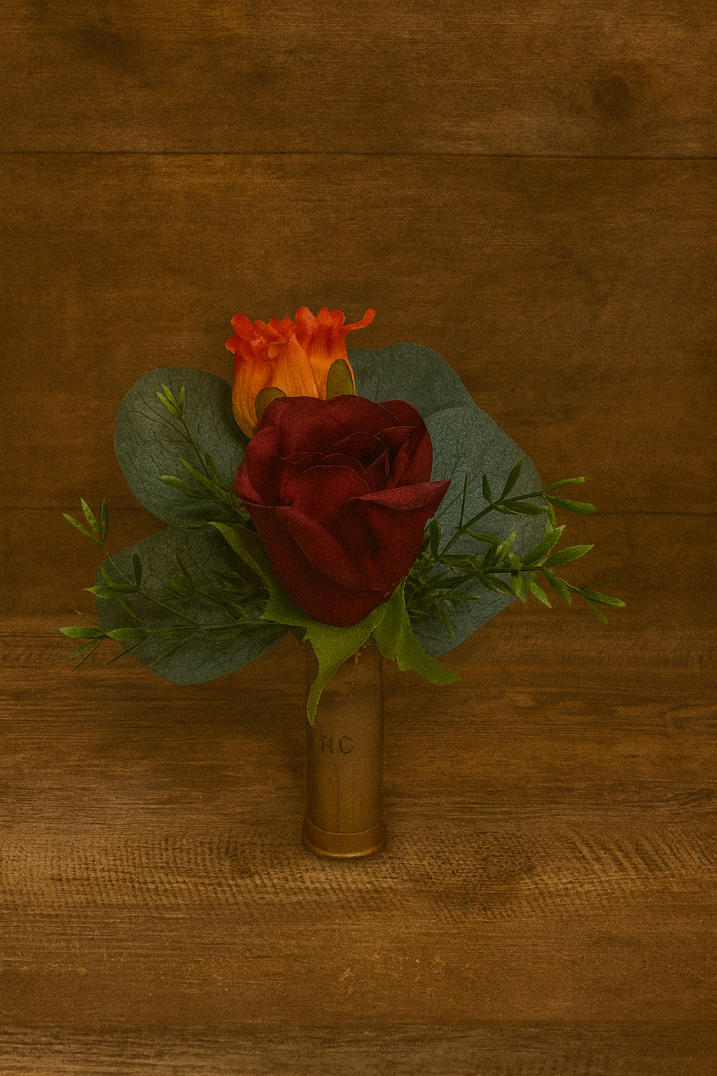 A boutonnière arrangement of a red rose, orange flower, and greenery, with a shotgun shell base, on a wooden surface.