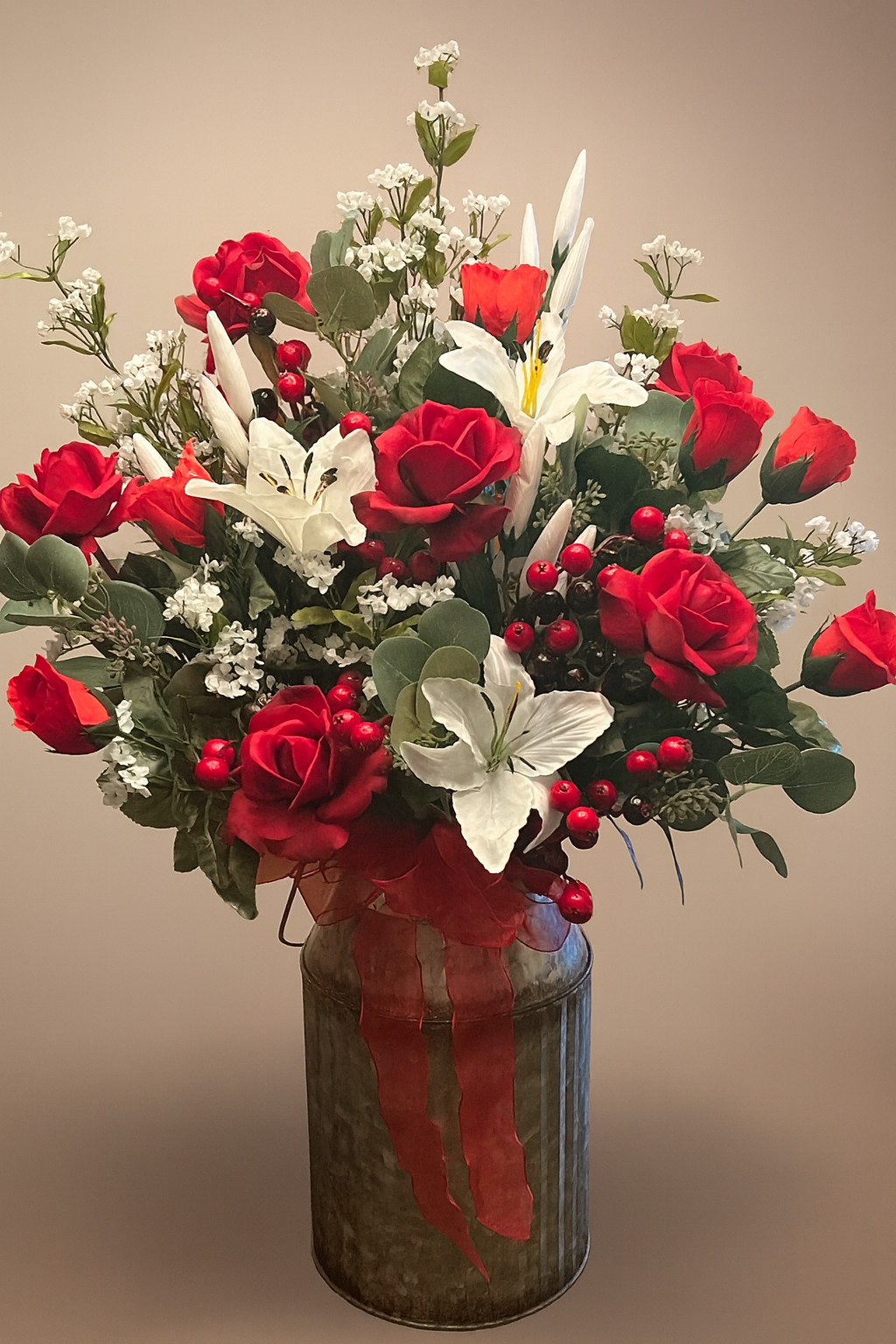 A floral arrangement with red roses, white lilies, baby's breath, and red berries in a metallic vase with a red ribbon.