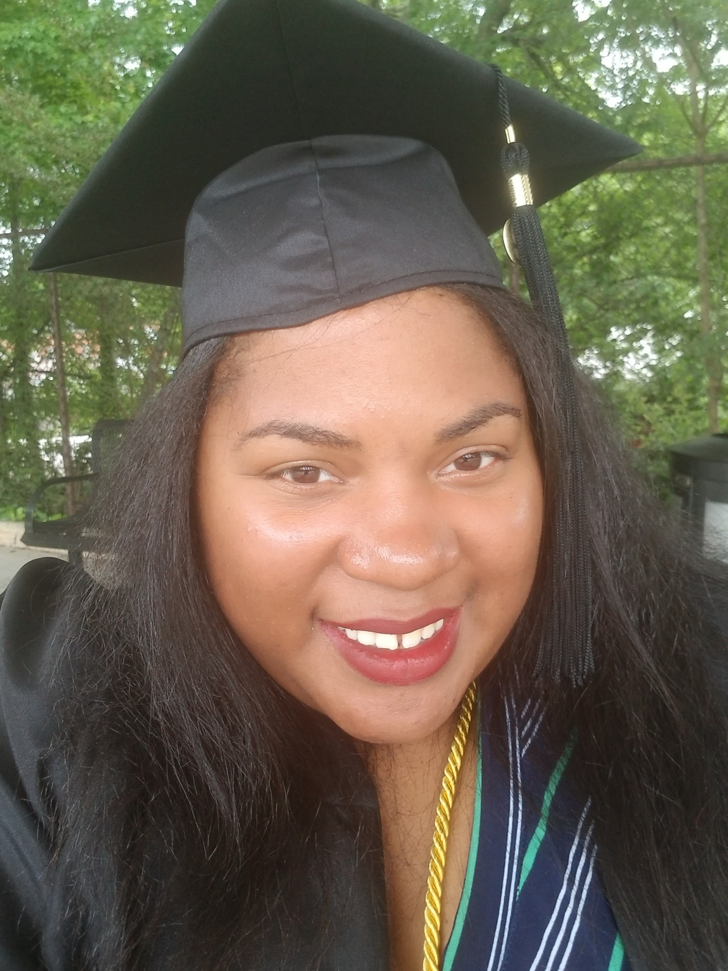 A woman in a graduation cap and gown smiling outdoors surrounded by green trees.