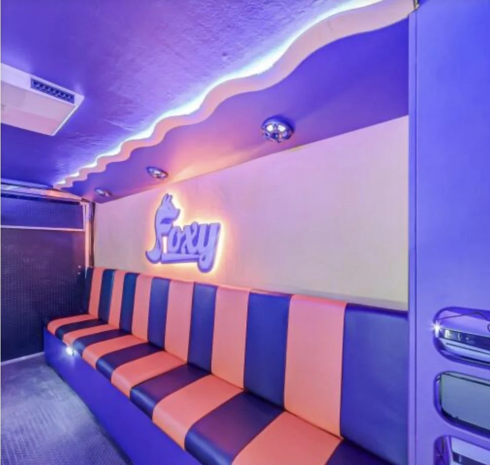 Retro-style lounge with a long striped orange and blue couch, neon sign with a fox and the word "Foxy," and purple and pink LED lighting accenting the ceiling.