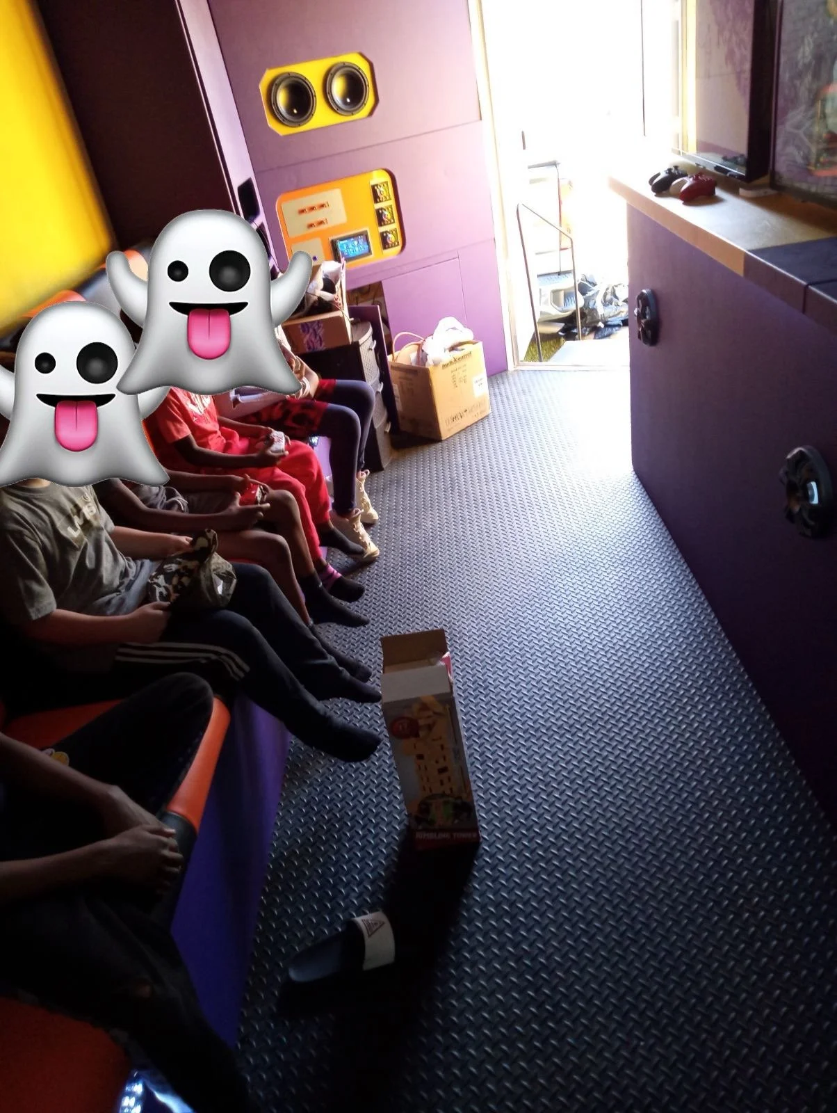 Children sitting on a colorful couch with purple walls, with a bright door open to sunlight outside. There are large speakers on the wall. In the foreground, there are some snack boxes and a bottle on the floor, and two ghost emojis overlay the image.