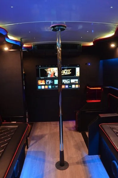 Inside a dimly lit party bus with a pole in the center, video screen on the back wall, and seating with red trim around the edges.
