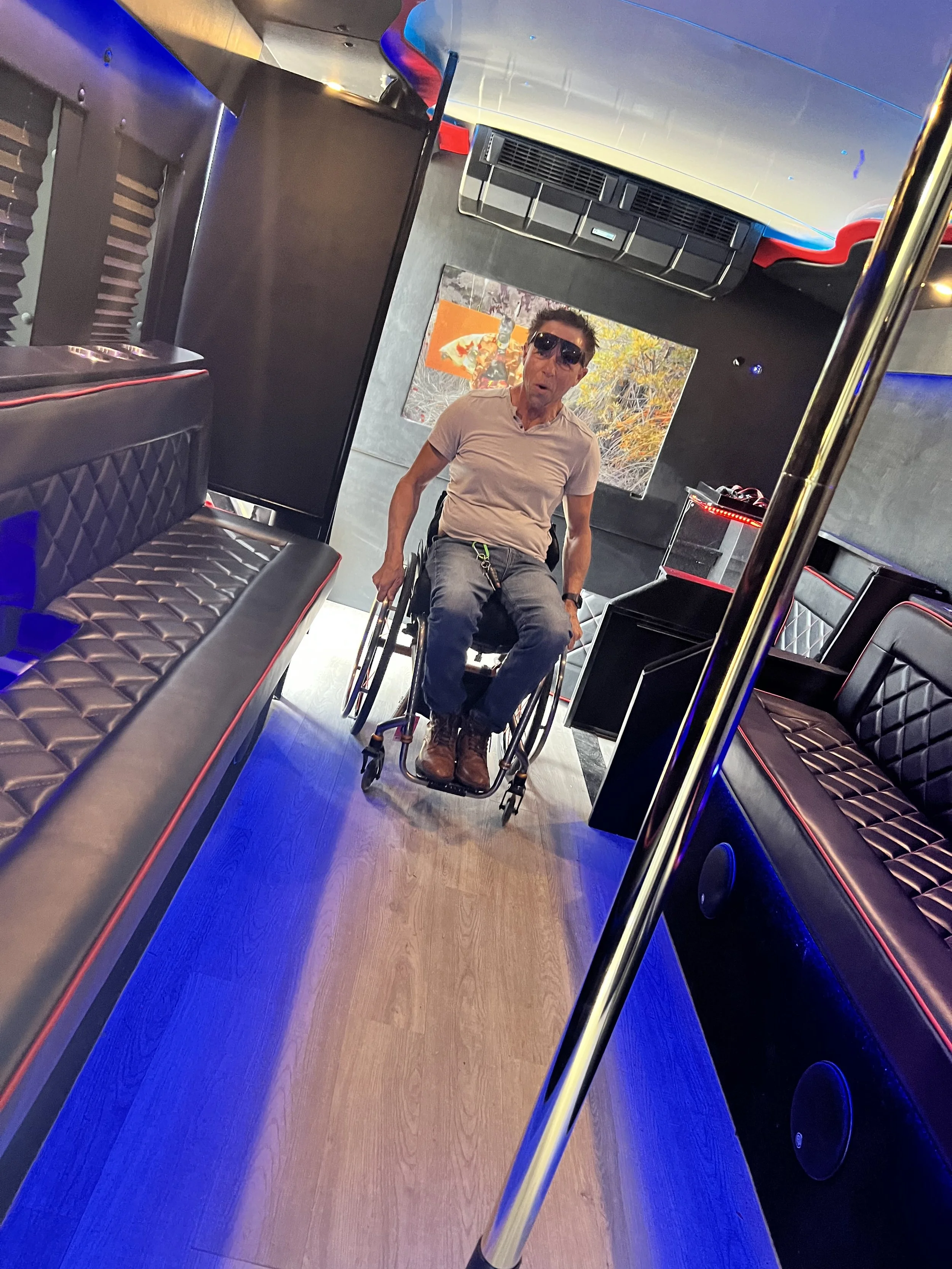 A man in a wheelchair inside a party bus with black leather seats and blue lighting, looking surprised or excited.