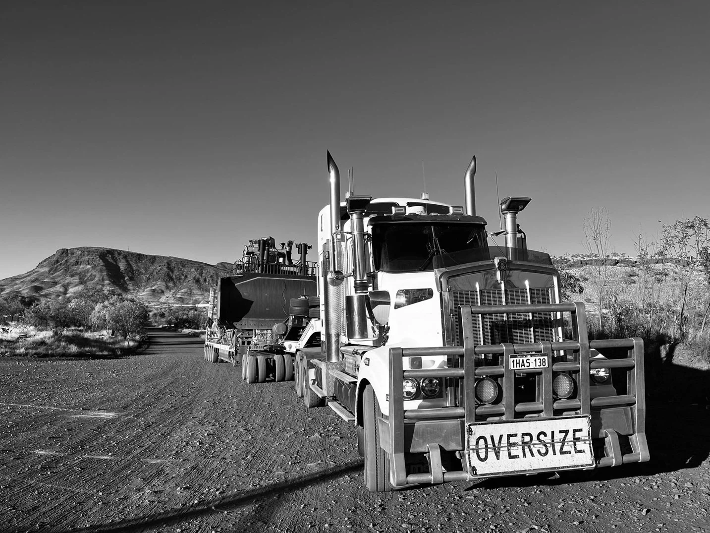 A large oversized semi-truck with a white cabin and a front bumper labeled 'OVERSIZE,' parked on a dirt road with hills and sparse vegetation in the background, in black and white.