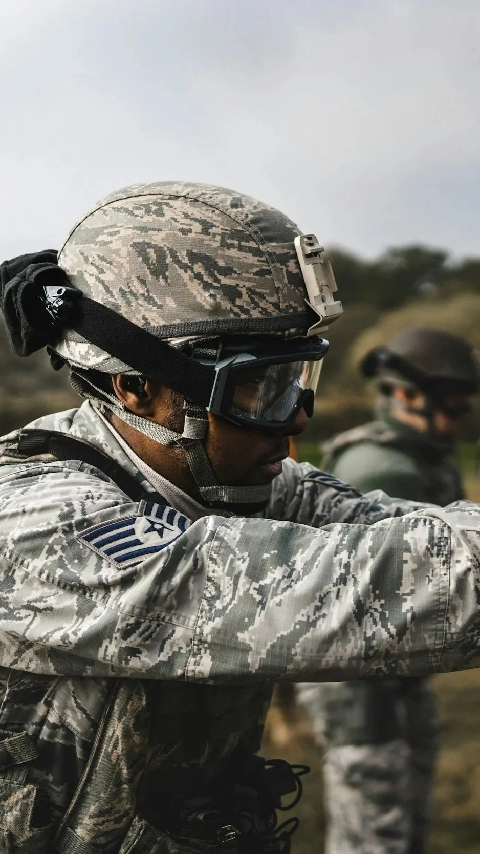 A soldier in camouflage military uniform wearing a helmet and protective goggles, with another soldier in the background.