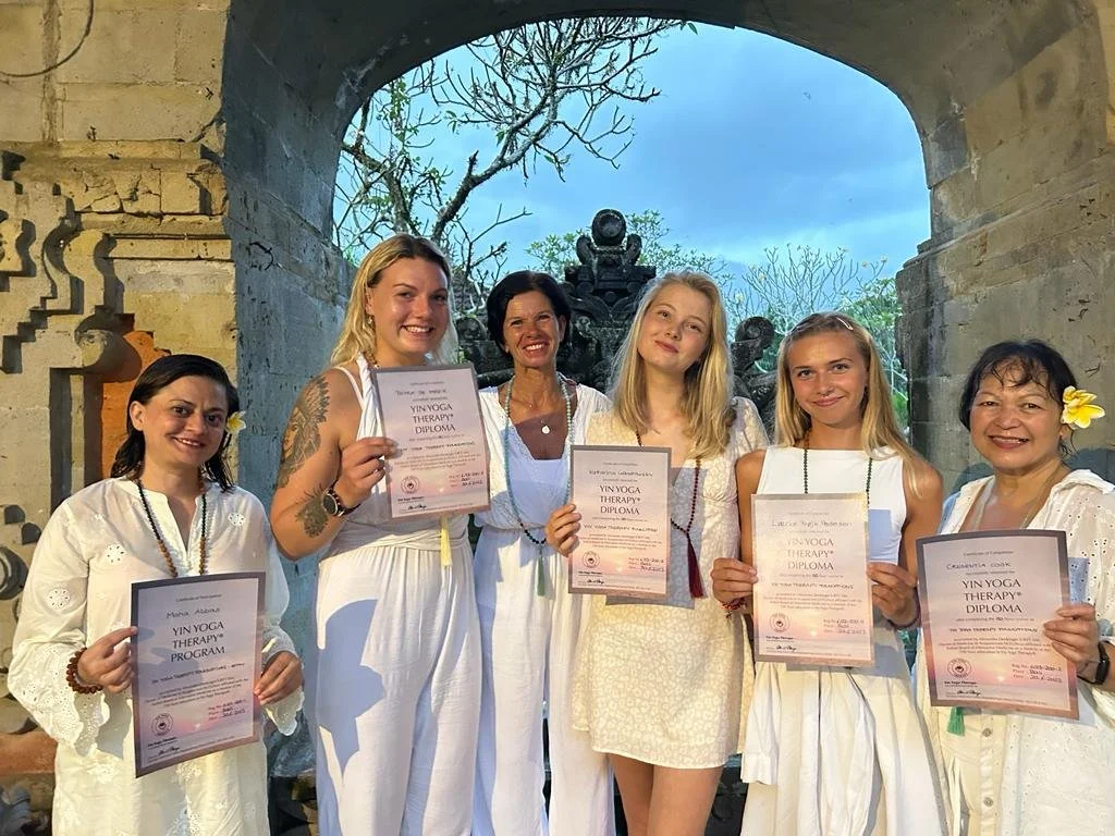 Group of six women standing together outdoors, holding yoga therapy diplomas, smiling, with a stone arch and trees in the background.