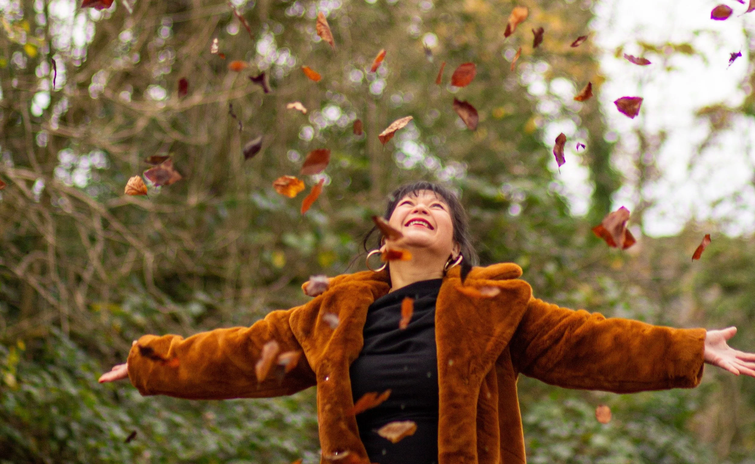 A woman in a brown coat with outstretched arms, smiling while autumn leaves fall around her in a forest.