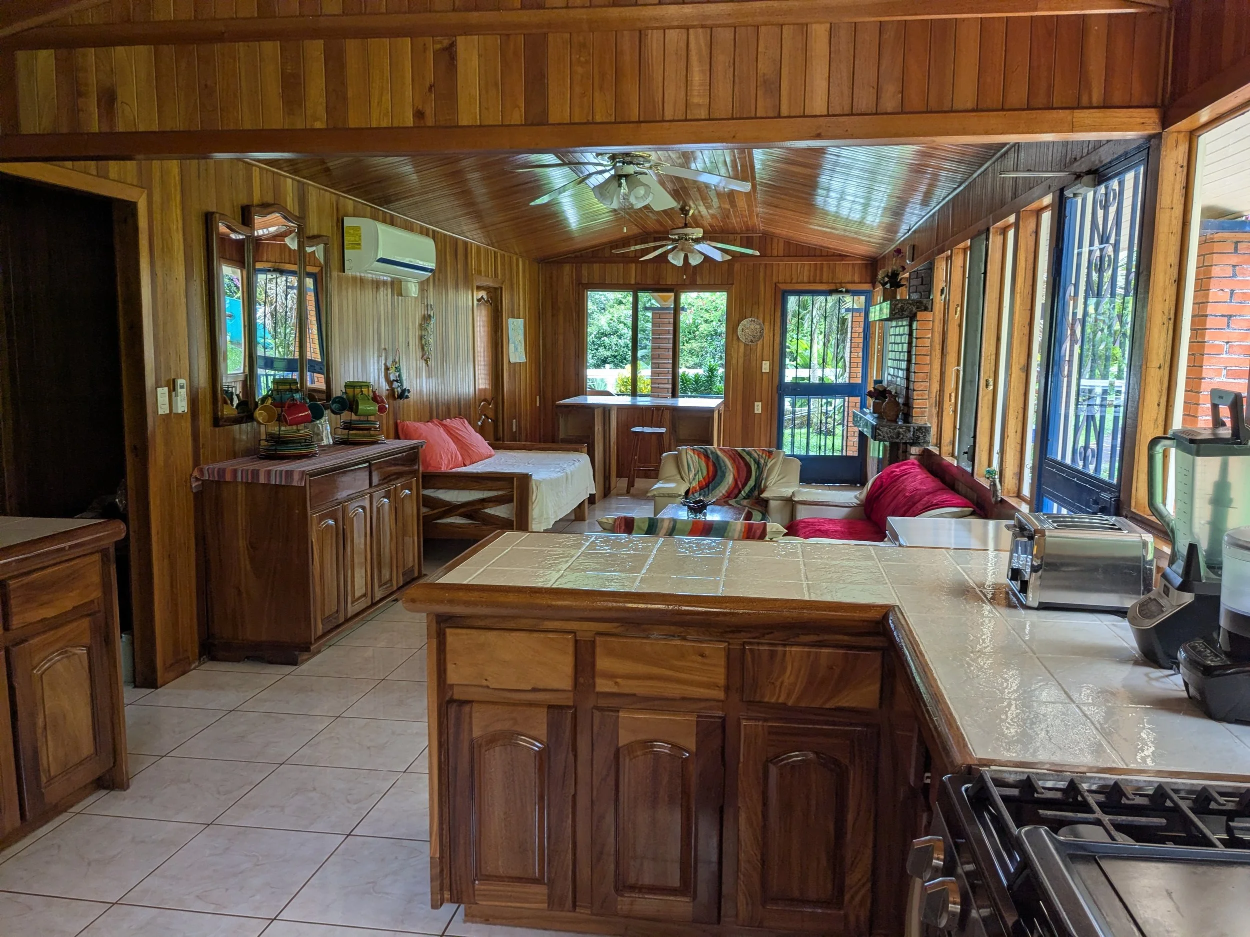 Interior of a wooden house with a kitchen area in the foreground, featuring a tile countertop, stove, and small appliances. In the background, a living room with couches, a coffee table, and large windows showing greenery outside.