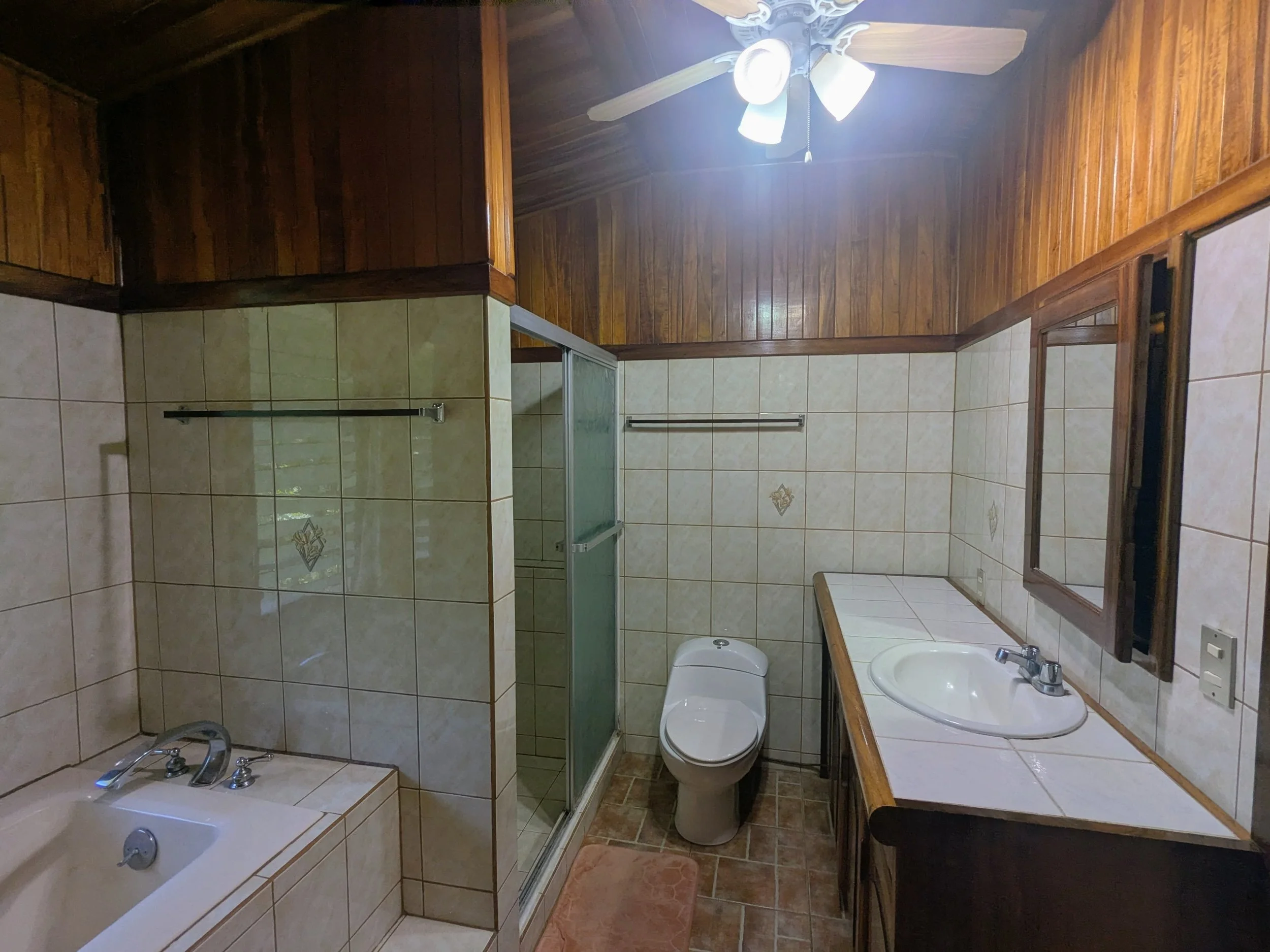 Bathroom with wood-paneled ceiling, beige tiled walls, a bathtub, a shower, a toilet, a sink with a mirror, and a ceiling fan with lights.
