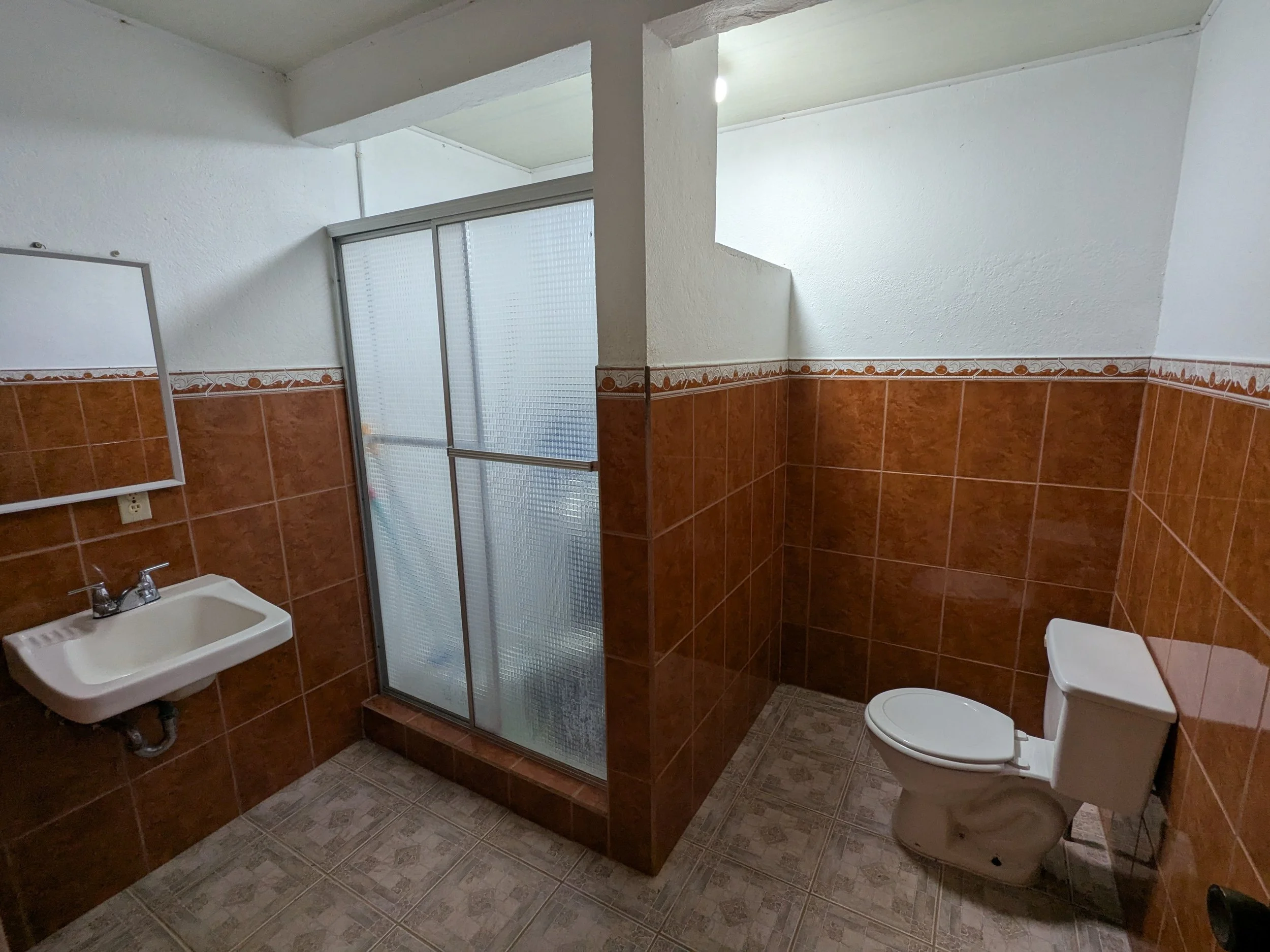 Bathroom with brown tiled walls, a small white sink with a chrome faucet, a frosted glass shower stall, and a white toilet.
