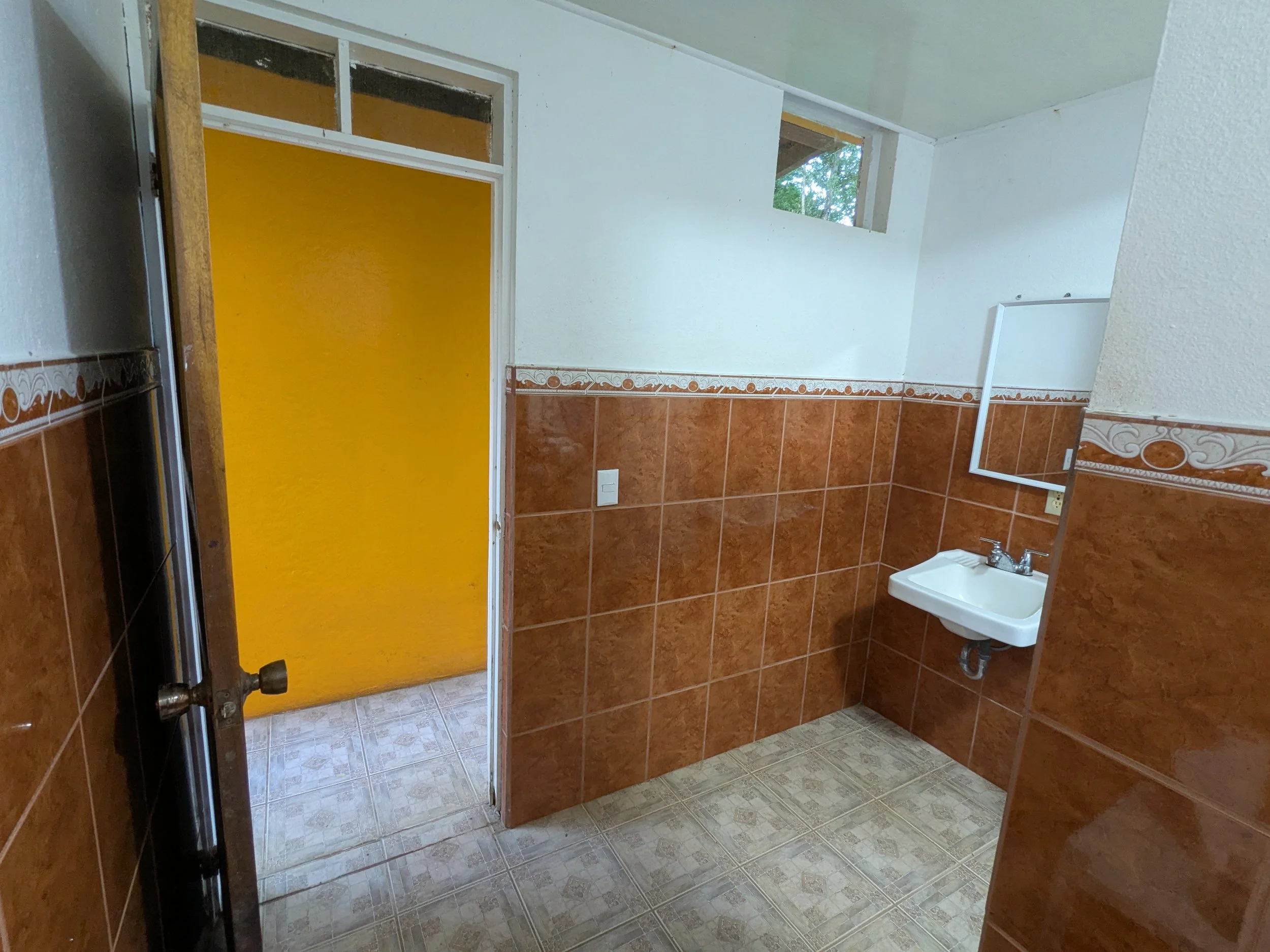 Small bathroom with brown tile walls, a white sink, and a mirror. The door has yellow paint on the other side. There are small windows near the ceiling letting in natural light.
