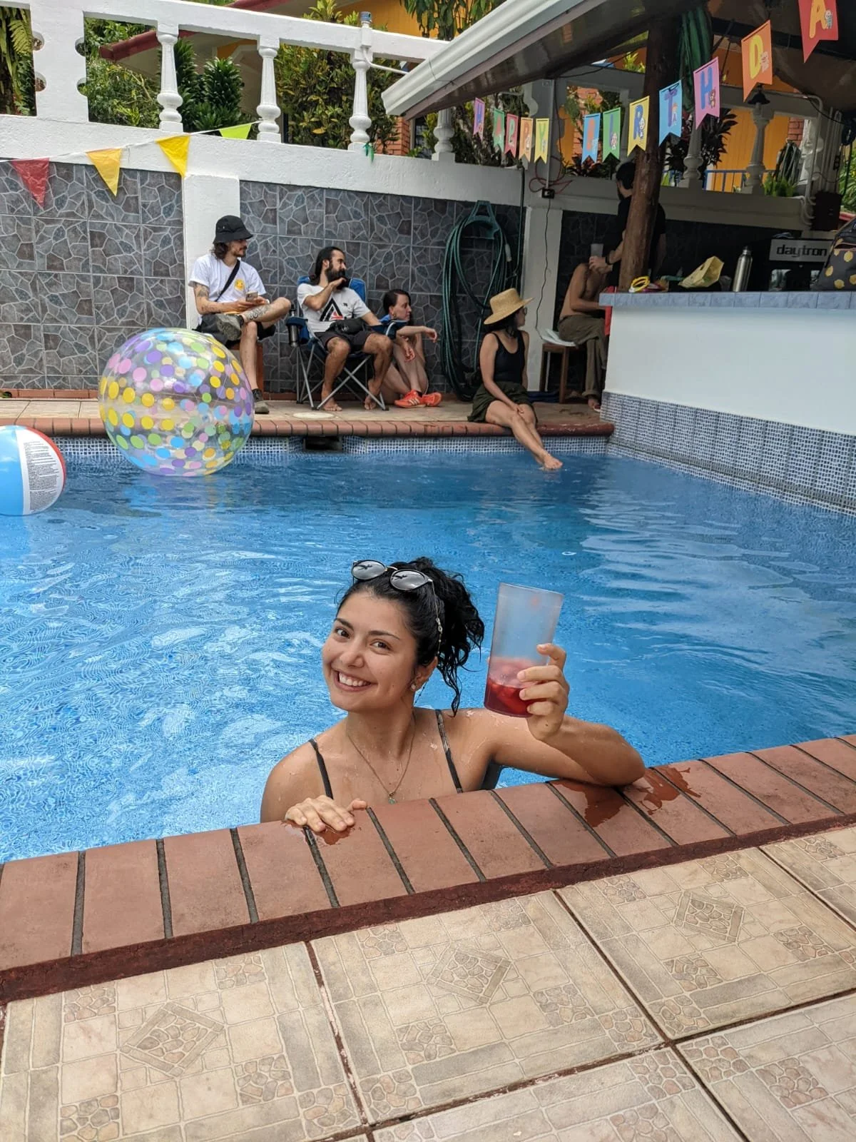 A young woman with sunglasses on her head smiling and holding a red drink in a clear plastic cup while in a swimming pool, with other people sitting on the poolside and chairs in the background, decorated with colorful banners and pool toys.