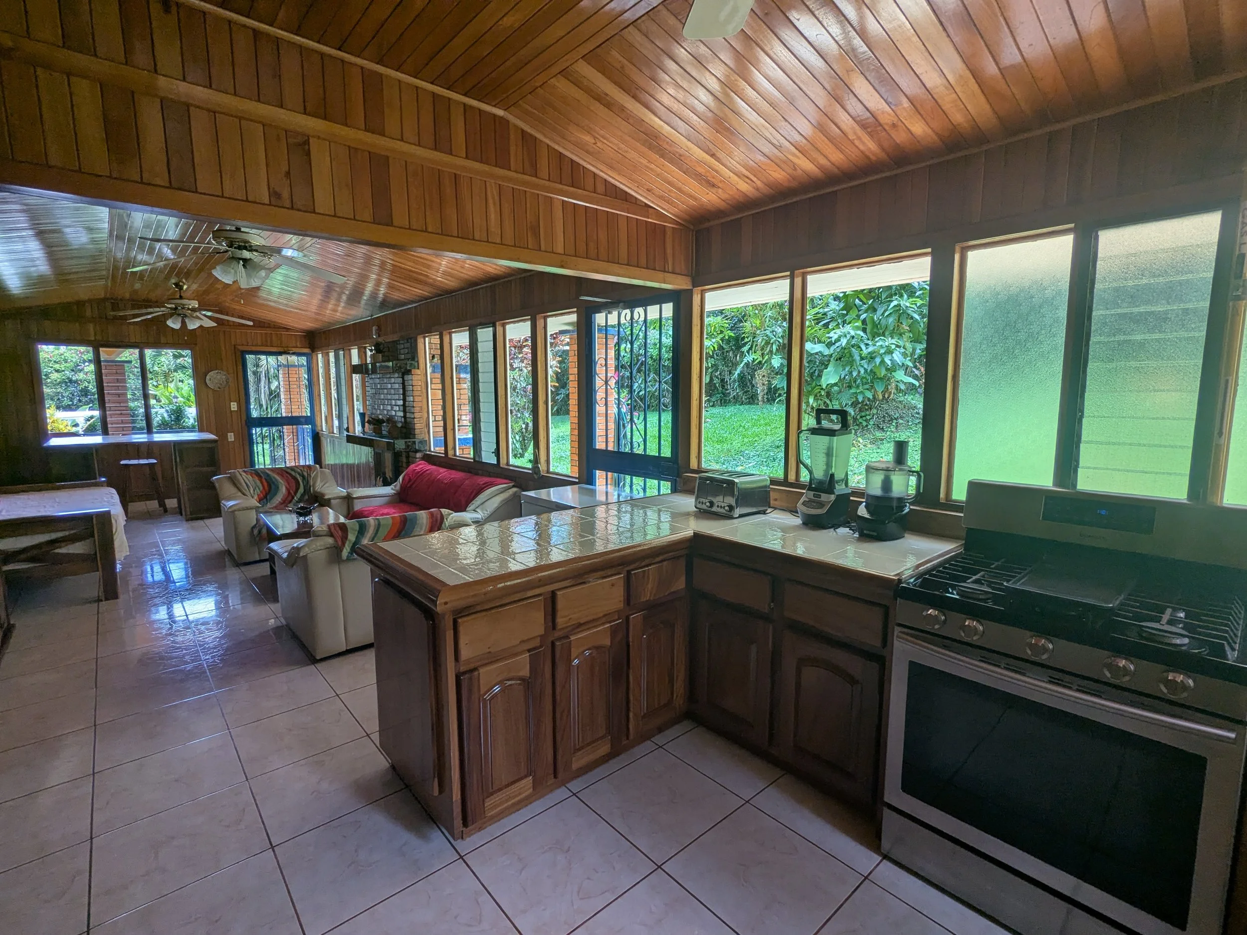 Kitchen and living room area with wood-paneled ceiling and walls, tiled floor, large windows showing green outdoor space, stove, countertops with small appliances, and white sofa with colorful blanket in the living room.