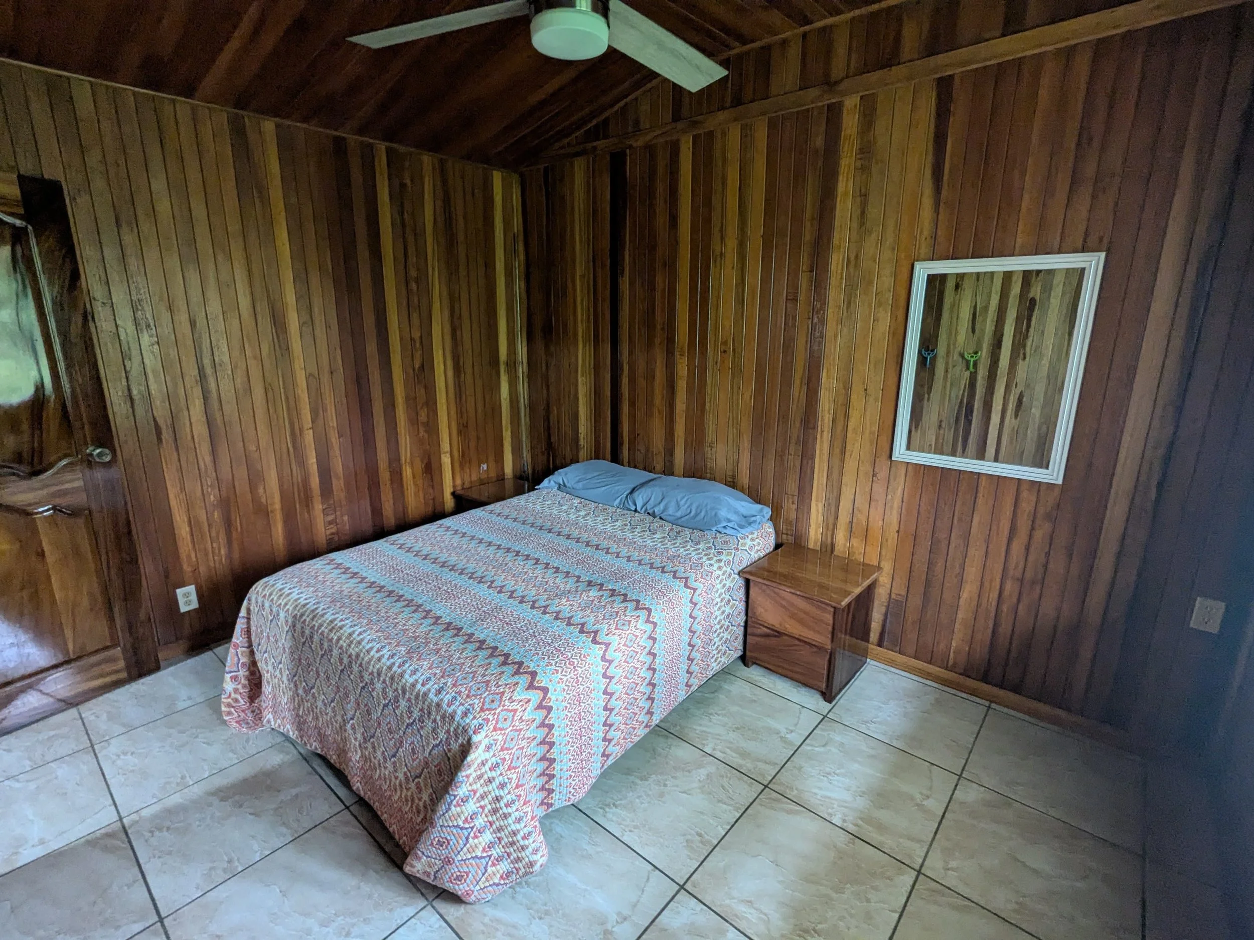 A bedroom with wooden paneled walls, a bed with a colorful patterned bedspread and two pillows, two small wooden nightstands, a white frame mirror on the wall, and tiled floor.