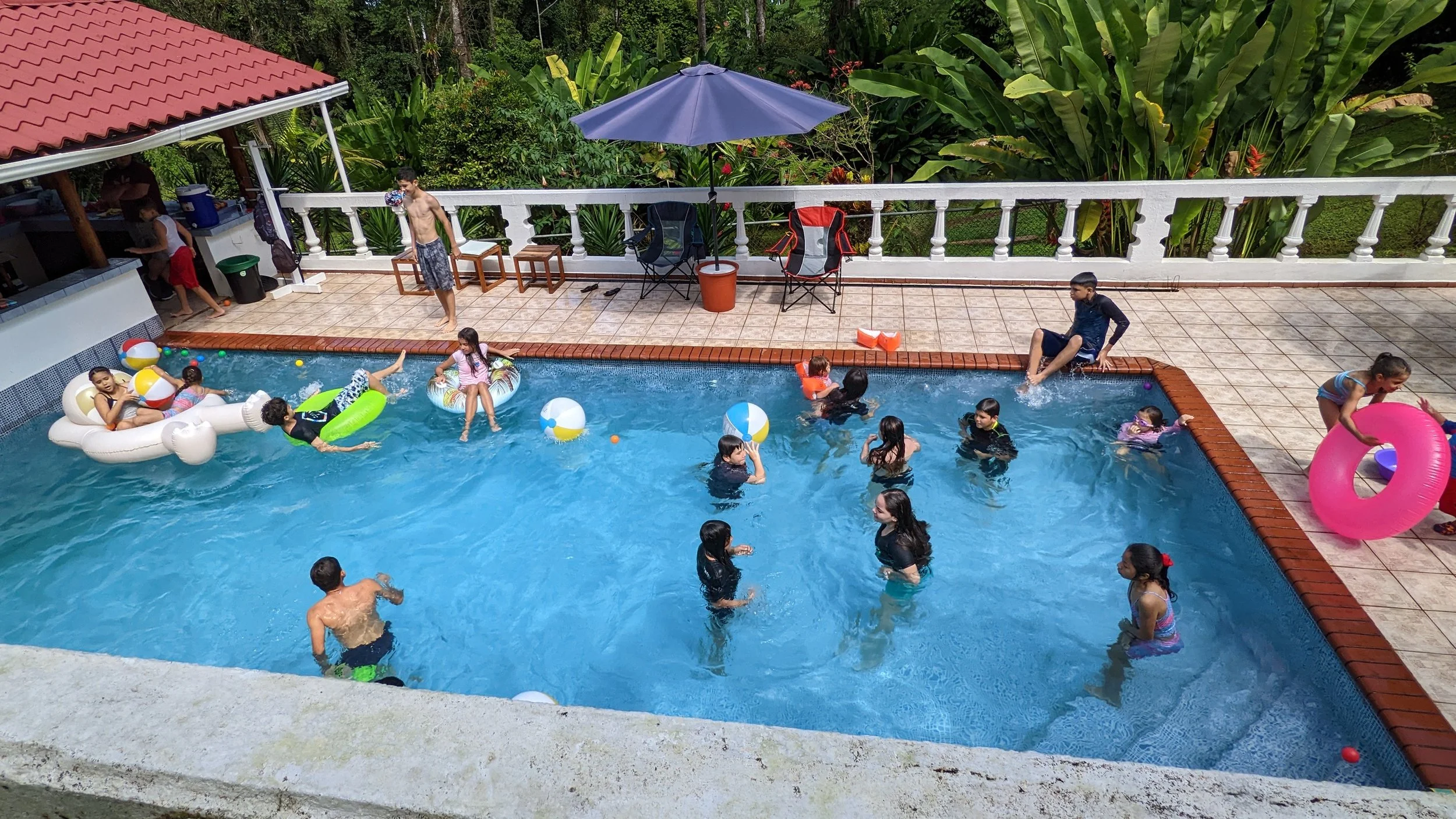 Children playing and swimming in a backyard pool, with inflatables and beach balls, surrounded by lush greenery and outdoor seating.