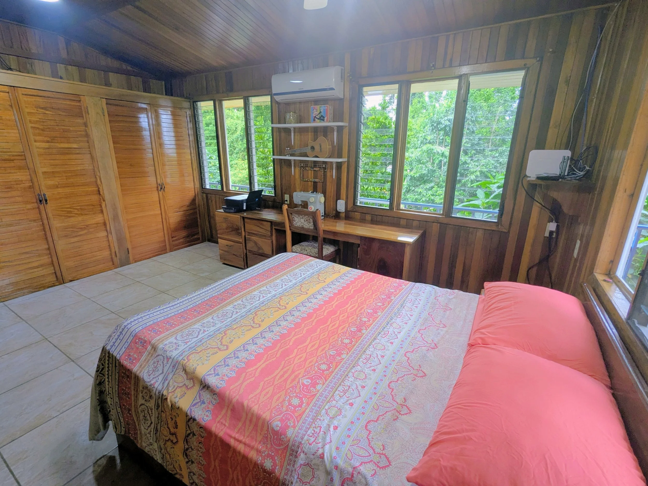 A cozy bedroom with wood-paneled walls and ceiling, large windows showing greenery outside, a bed with a colorful patterned bedspread and pink pillows, a wooden desk with a sewing machine and printer, and closed wooden closet doors.