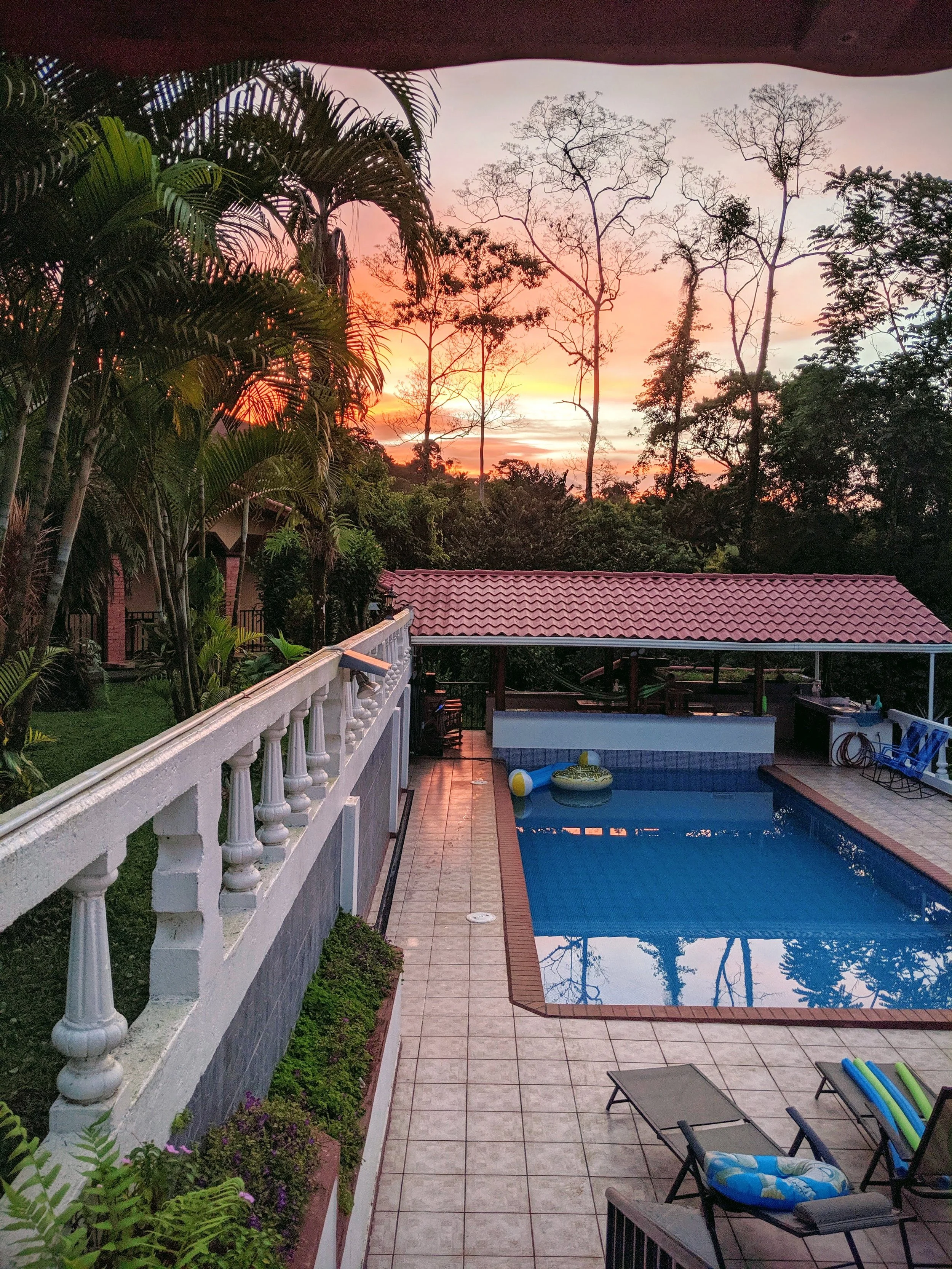 A backyard swimming pool area during sunset with lounge chairs, pool floaties, a barbecue area, and surrounding trees with a pink and orange sky.